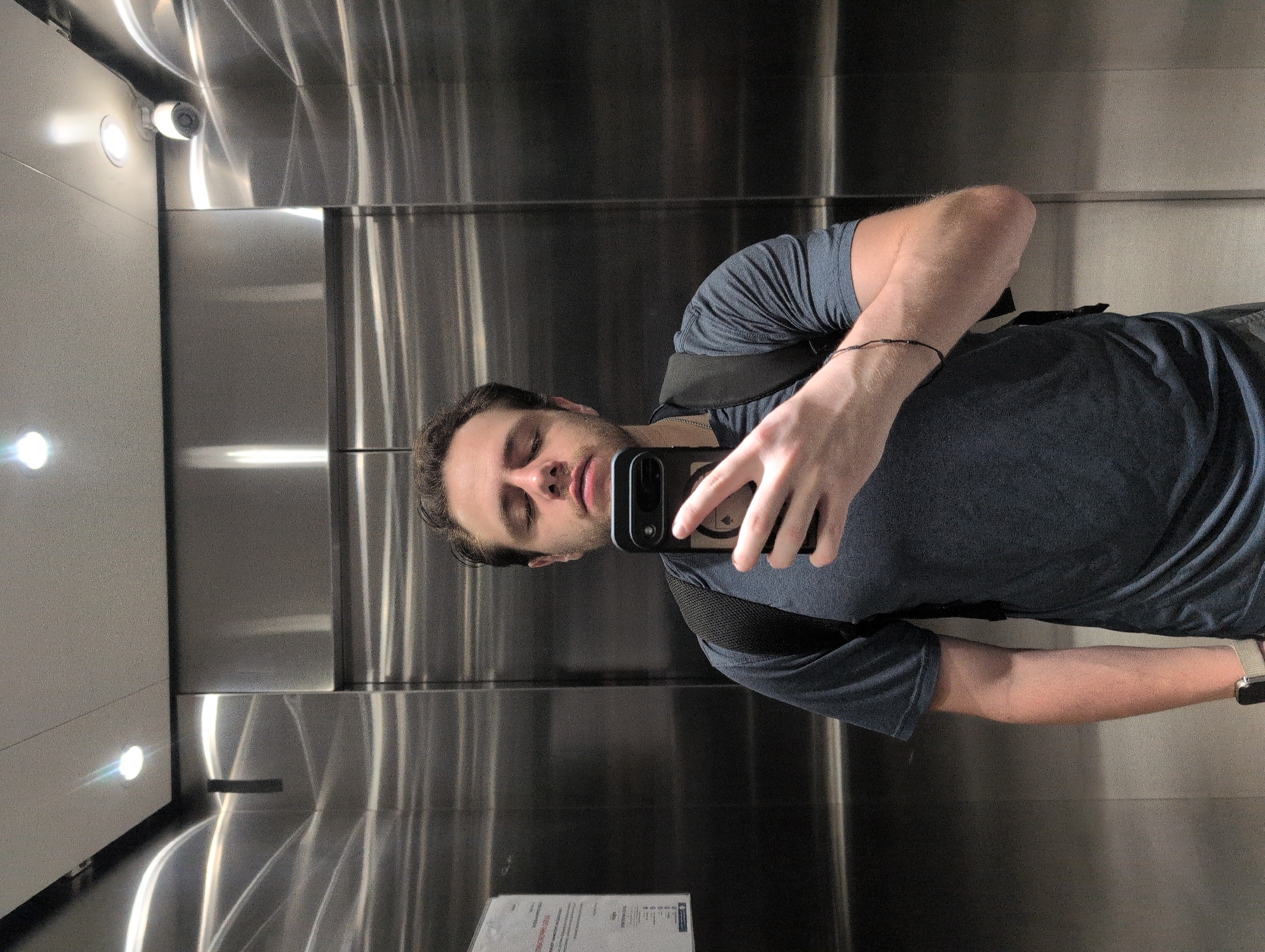 A man is taking a selfie in what appears to be an elevator. The elevator has metal walls with visible seams and lighting fixtures. A security camera is visible in the upper left corner. A piece of paper with text is visible at the bottom of the image.