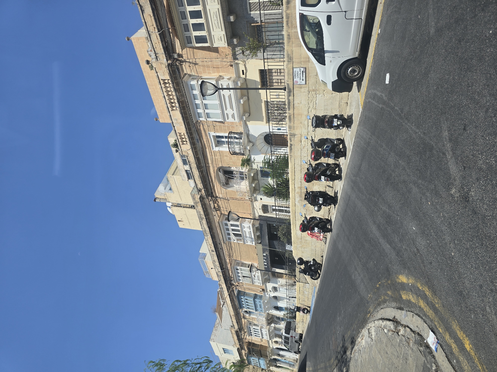 The image shows a street in Saint Julian's, Malta. The street slopes downward from left to right. On the right side of the street, there is a white van parked. Further down the street, there are several parked scooters. A wall runs along the right side of the street, behind the scooters. On top of the wall, there are several residential buildings with balconies. The buildings are light brown and have ornate architectural details. The sky is clear and blue. The image appears to have been taken on a sunny day.
