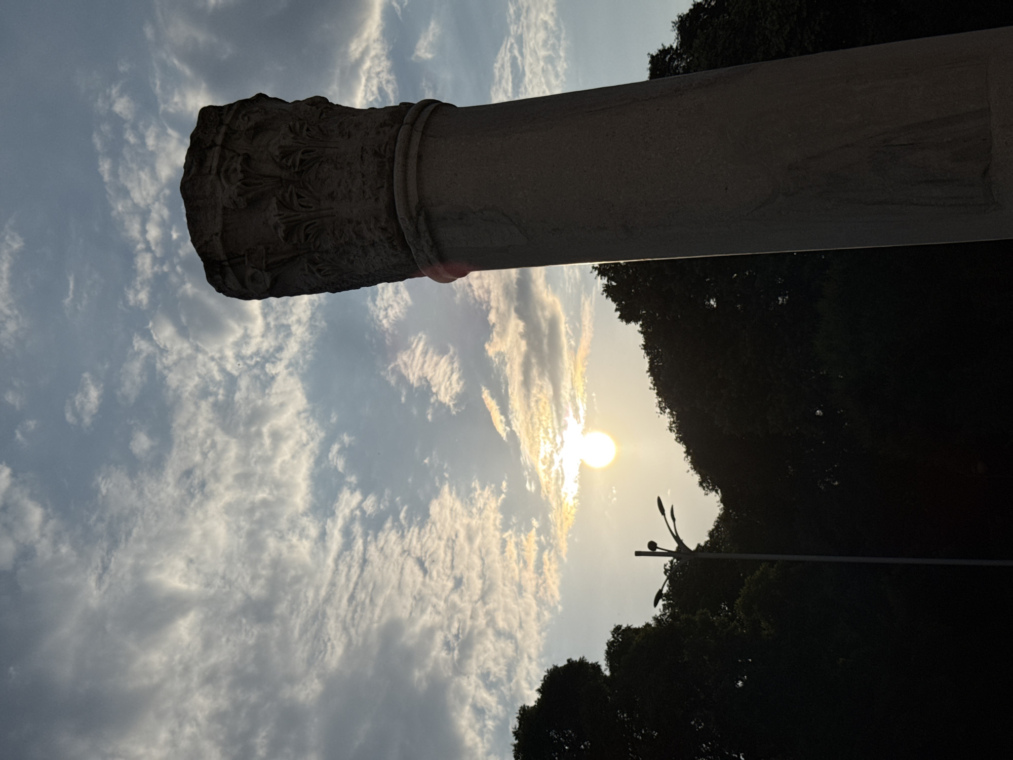 The image displays an outdoor scene captured during either sunrise or sunset, characterized by strong backlighting. A large, weathered stone column occupies the right side of the frame, extending from the bottom to near the top, rendered in silhouette. Its capital is intricately carved with stylized leaf motifs, indicative of a classical architectural order, possibly Corinthian. The column's shaft is smooth and appears to be made of stone.
The sun, a bright, circular, yellowish-white orb, is prominently positioned in the middle-left, low on the horizon, partially obscured by light clouds. Its intense light creates a lens flare visible as a faint reddish streak along the inner edge of the column. The sky above is a mosaic of pale blue, white, and light grey clouds, with some clouds near the sun illuminated with warm, golden hues.
Below the horizon line, dense, dark green and black foliage, consistent with a cluster of trees, fills the lower left and bottom central portions of the image, appearing in deep silhouette. A modern, tall streetlamp with multiple arm-mounted lights is silhouetted against the brighter sky, positioned between the trees and the sun. No human figures or animals are present in the scene.
This column is identified as a feature within the Roman Forum complex of Plovdiv, Bulgaria, a city renowned for its preserved ancient Roman architecture.