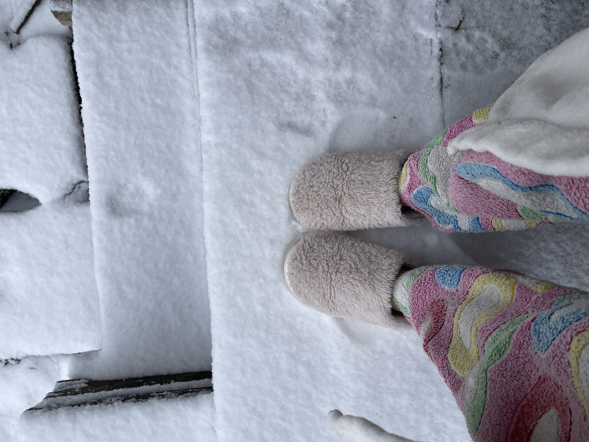 A top-down, full shot depicts a pair of feet clad in light-colored, fuzzy slippers and patterned pajamas, standing on snow-covered wooden steps. The steps are heavily blanketed in snow, obscuring the wood grain and edges. The pajamas feature a vibrant, abstract pattern with pink, yellow, green, blue, and white hues. A portion of a white robe is also visible. The photograph's composition is simple and focuses on the subject's feet in the snowy environment. There is no indication that this is in the city of Фоки, Russia.