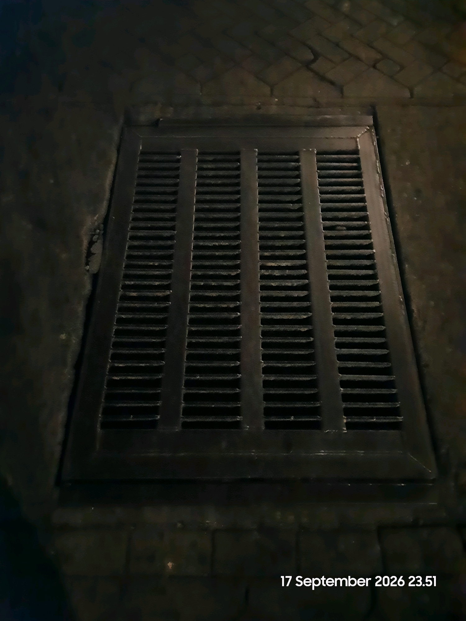 The image shows a close-up, eye-level view of a dark metal grate set into a dark, textured surface that appears to be pavement or cobblestone. The grate is rectangular and composed of horizontal slats separated by vertical supports, dividing it into four sections. The surface surrounding the grate is uneven and appears to be dimly lit. A timestamp, "17 September 2026 23.51," is visible in the lower right corner of the image. The scene is devoid of any people or discernible activity. The context suggests an urban or semi-urban environment, likely outdoors at night. There is no information within the image itself to specify the location as Gending, Indonesia.