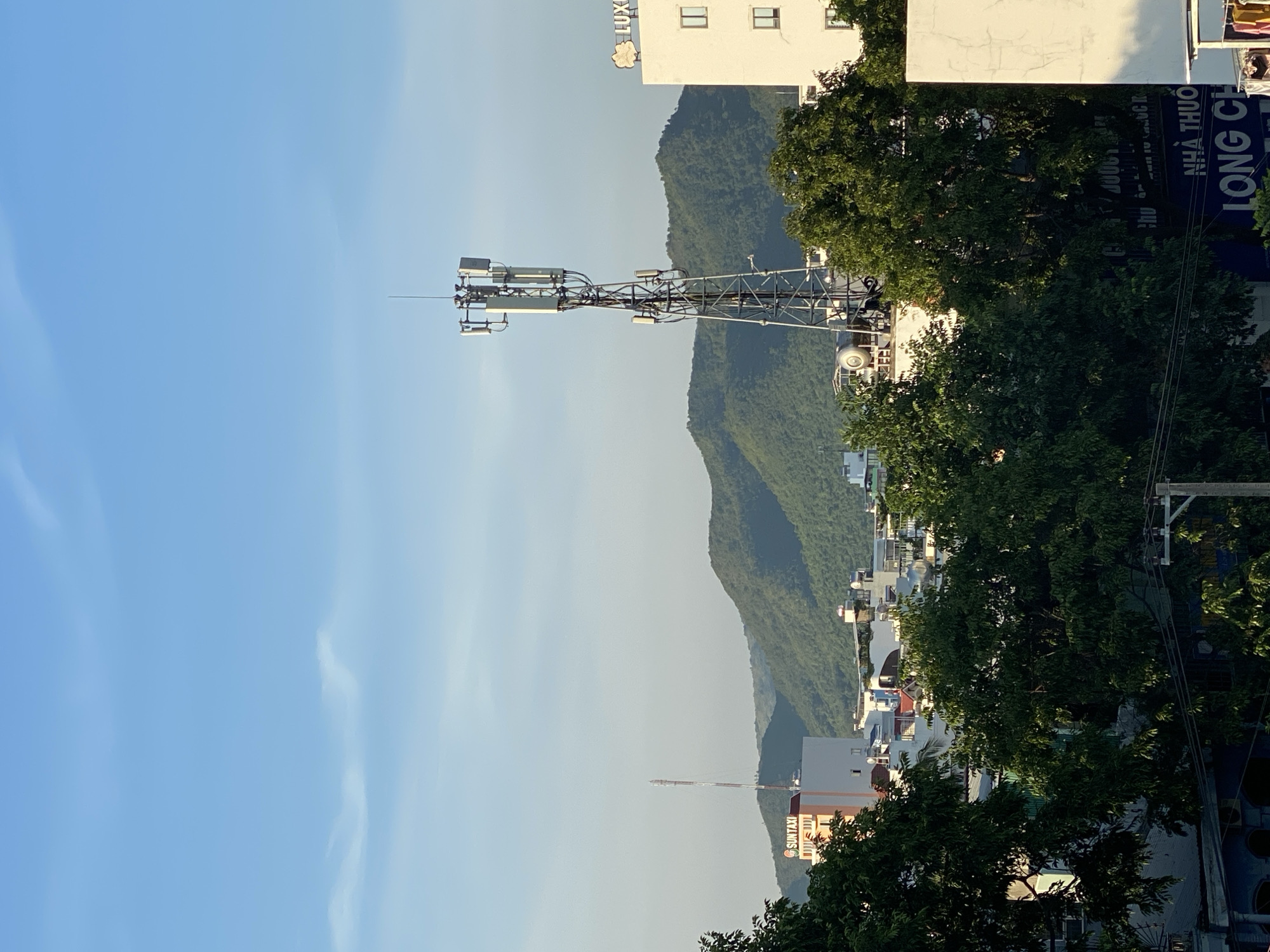 The image captures a cityscape with a backdrop of lush green mountains under a clear blue sky. In the foreground, dense green trees partially obscure a cluster of buildings. A tall communication tower stands prominently in the middle ground, adorned with various antennas and equipment. To the right of the tower, a white building with visible windows is present, and a portion of a sign reading "LUX..." is discernible. To the left of the tower, another building features a sign that says "SUN TAXI." The overall scene depicts an urban environment interspersed with natural elements.