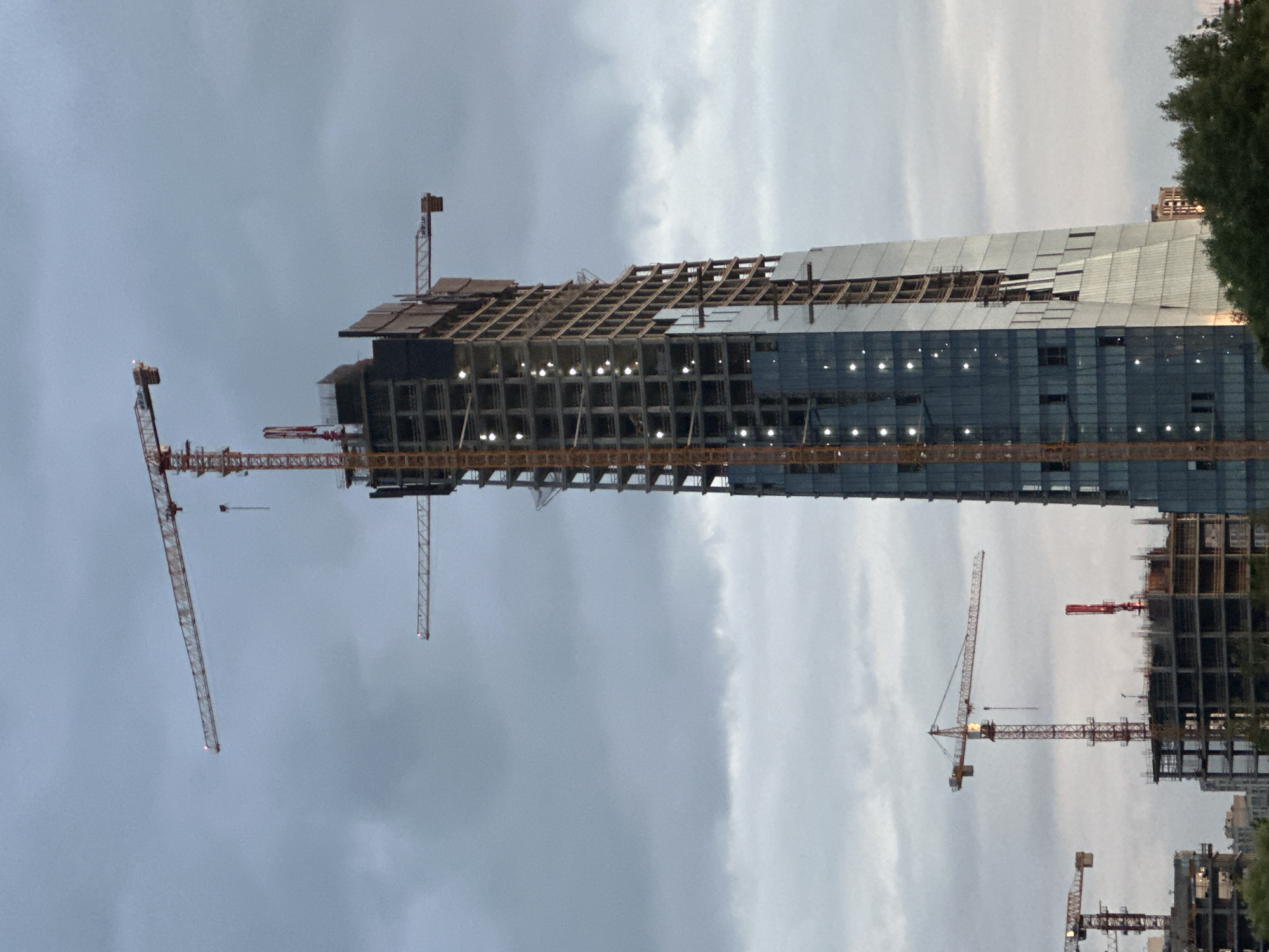 An upward-angled, wide shot captures a cluster of active construction sites under an overcast, diffused grey sky, likely at dusk or dawn. Dominating the frame is a towering, slender high-rise building in the center-right, still under construction. Its lower half is largely clad in dark, reflective glass panels, while the upper half reveals the exposed concrete framework with numerous interior lights illuminating the structure. The building exhibits a distinct architectural design with a subtle inward curve or taper on its right side.
Two large tower cranes are prominently associated with this main building: an exceptionally tall crane with its jib extending far to the left, and a smaller crane jib extending horizontally from the building's right side. In the mid-ground, to the left of the main high-rise, another freestanding tower crane with a red mast is visible, actively engaged in construction over a partially built concrete structure. A bright red concrete pump is also visible near this mid-ground crane. Further left, parts of additional construction cranes and lower-rise concrete building frames are visible, indicating extensive urban development. Green foliage is present in the lower right foreground. The scene is located in the city of Minsk, Belarus.