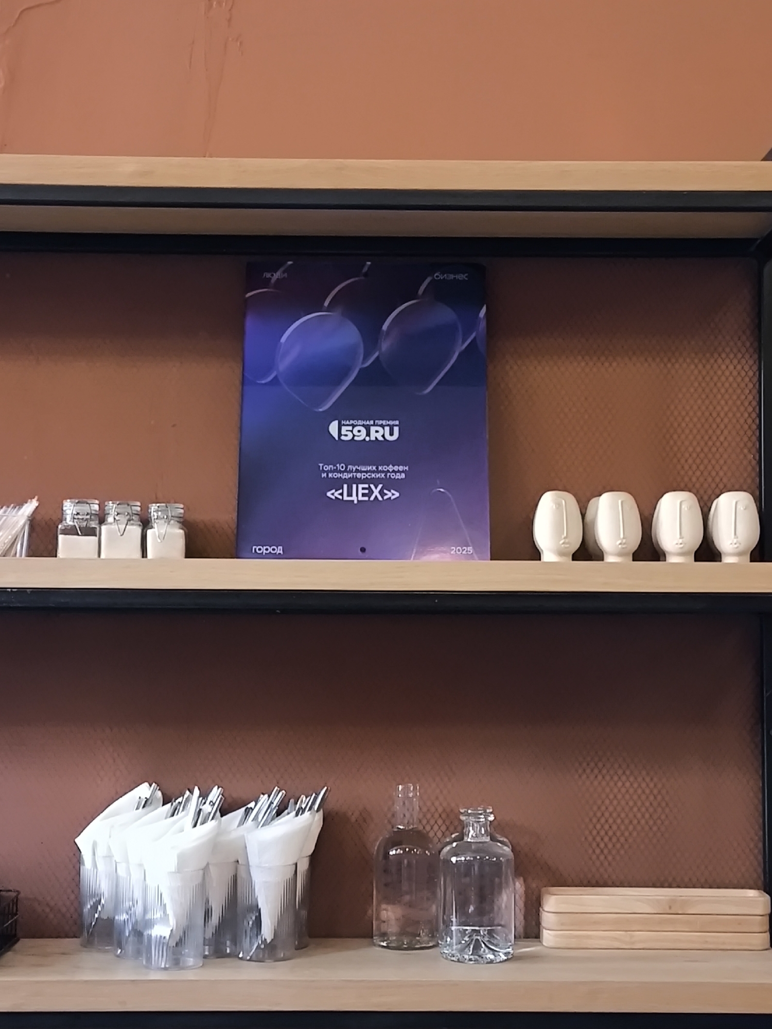 The image shows a shelf in what appears to be a cafe or restaurant. On the shelf, from left to right, are three small glass jars with metal lids, each containing a white substance. Next to them is a poster with a dark blue background featuring two abstract purple shapes and text in Russian. The text reads "НАРОДНАЯ ПРЕМИЯ 59.RU" (People's Prize 59.RU), "Топ-10 лучших кофеен и кондитерских года" (Top 10 best coffee houses and confectioneries of the year), and "<<ЦЕХ>>" (Workshop) with the year "2025" at the bottom. To the right of the poster are five white ceramic objects shaped like stylized human faces.
The second shelf, visible in the lower part of the image, holds several clear plastic cups filled with napkins and cutlery. Further to the right are two clear glass bottles, one tall and slender, the other shorter and wider with a textured base. To the far right are three stacked rectangular wooden trays.
The background wall is a textured reddish-brown color. The shelves are made of light-colored wood with black metal supports. The overall impression is of a clean, organized space, possibly a service area for customers. The poster references Perm, Russia, suggesting this establishment is located in that city.