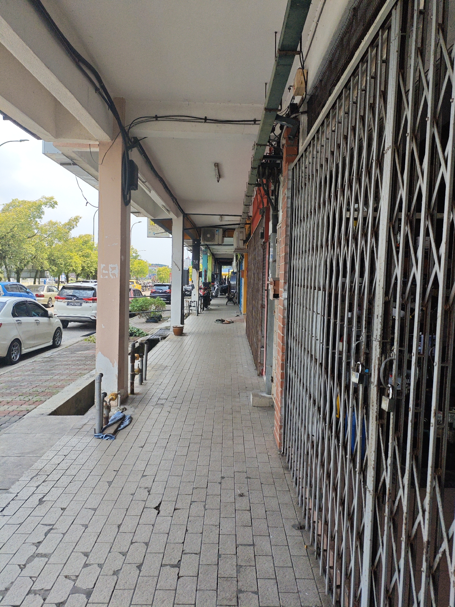 The image captures a street view in Kota Tinggi, Malaysia, showing a covered walkway in front of shop lots. The walkway is paved with light-colored square tiles, some of which appear worn or broken. On the left side, a row of parked cars, including a white sedan and a white SUV with license plate JTG 7620, are visible along the street. Trees with green foliage line the background. The walkway is supported by square, light-colored pillars. On the right side of the walkway, metal roller shutters, some with padlocks, are visible in front of closed shop entrances. Electrical wires and conduits run along the underside of the covered ceiling. A few potted plants and a brown ceramic pot are placed along the walkway. In the distance, more shop fronts and parked motorcycles can be seen. The overall scene suggests a commercial area, possibly during daytime, with businesses closed or yet to open.