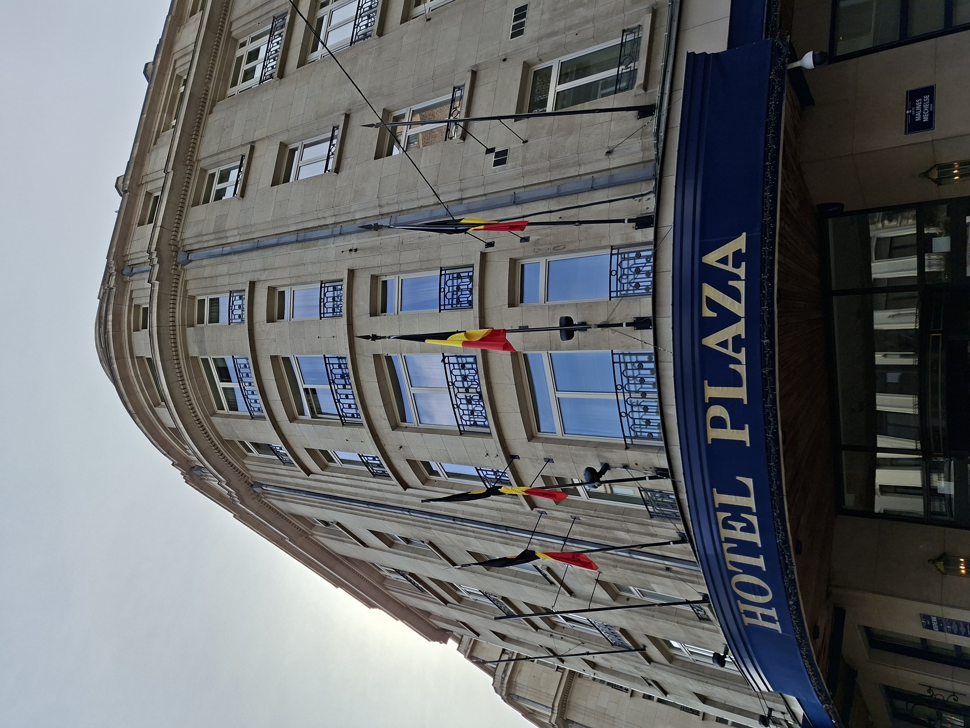 The image shows the facade of the Hotel Plaza, a building in Brussels, Belgium. The hotel's name is prominently displayed in gold lettering on a dark blue awning above the entrance. The building is made of light-colored stone and has multiple stories with numerous windows, many of which have decorative black metal balconies. Several Belgian flags are visible, mounted on poles extending from the facade. The sky is overcast, with the sun peeking through on the left side of the image, casting a slight glare. The architecture appears to be classical or Beaux-Arts style. The perspective is low-angle, looking up at the building. There are no people visible in the image.