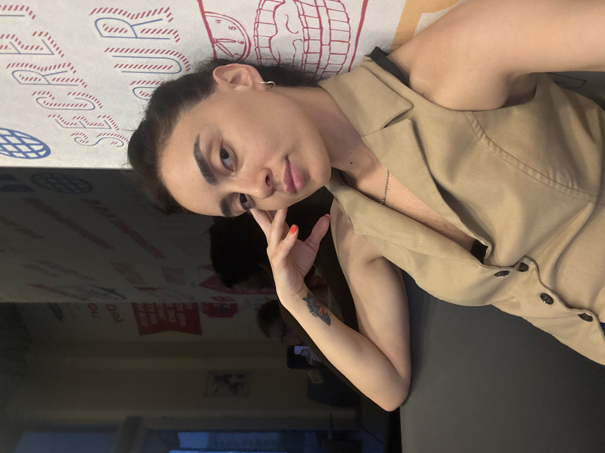 A woman is captured in a selfie-style photograph, taken at a slight angle from above. She has dark hair pulled back and is wearing a light brown sleeveless blazer over a dark top. Her left arm is resting on her lap, and she is looking towards the camera with a neutral expression. Her left hand is near her face, with bright orange nail polish visible. A small butterfly tattoo is present on her left forearm. A thin silver chain necklace is visible around her neck. The background appears to be a patterned surface, possibly a tablecloth or wallpaper, with red and blue designs. The overall lighting suggests it is daytime. The setting is indoors, possibly in a home or a studio, given the reflective surface in the foreground that shows a glimpse of other objects and possibly a phone screen. The location is identified as Tbilisi, Georgia.