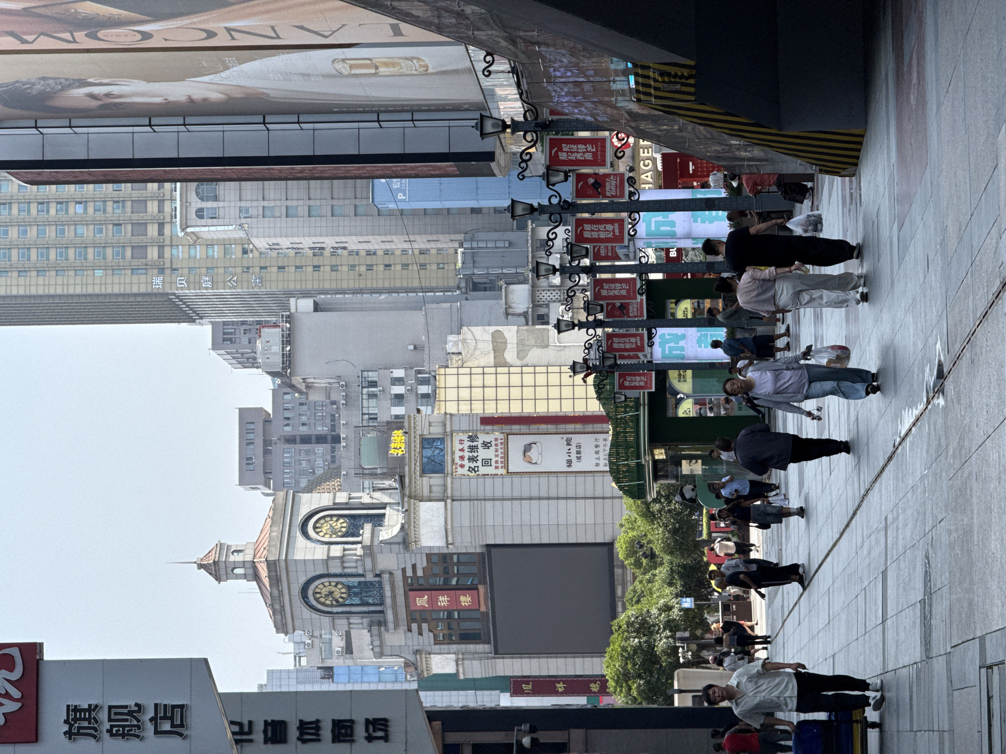 The image depicts a street scene in Jinjiang District, Chengdu, China. People are walking on a wide sidewalk. Various buildings line the street, including one with a clock tower and Chinese signage. There are numerous advertisements, some for luxury brands like Lancome and others with Chinese characters. Lampposts with ornate detailing are visible along the sidewalk. Overall, the scene appears to be a busy commercial area in a modern city.