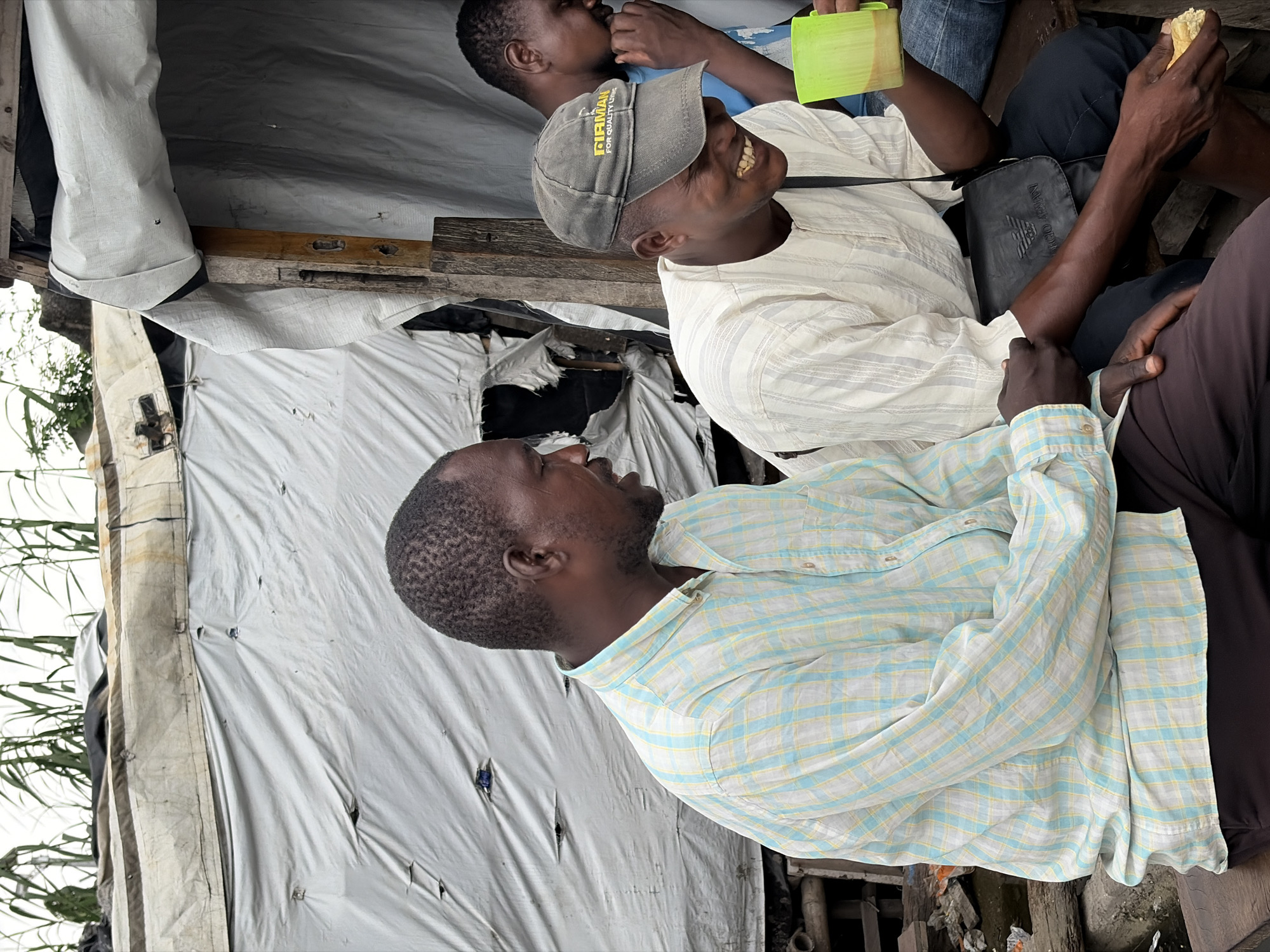 In Igboefon, Nigeria, two men are seated outdoors under a makeshift shelter constructed of tarp and wood. The man on the left, wearing a light blue and yellow plaid shirt, is looking to his right and speaking. The man on the right, wearing a light-colored striped shirt and a dark baseball cap, is laughing and looking down while holding a yellow cup with a brown liquid in his right hand and a piece of food in his left. A third man is partially visible in the background, wearing a blue shirt and touching his face. The environment appears to be rustic, with visible debris and rough wooden structures.