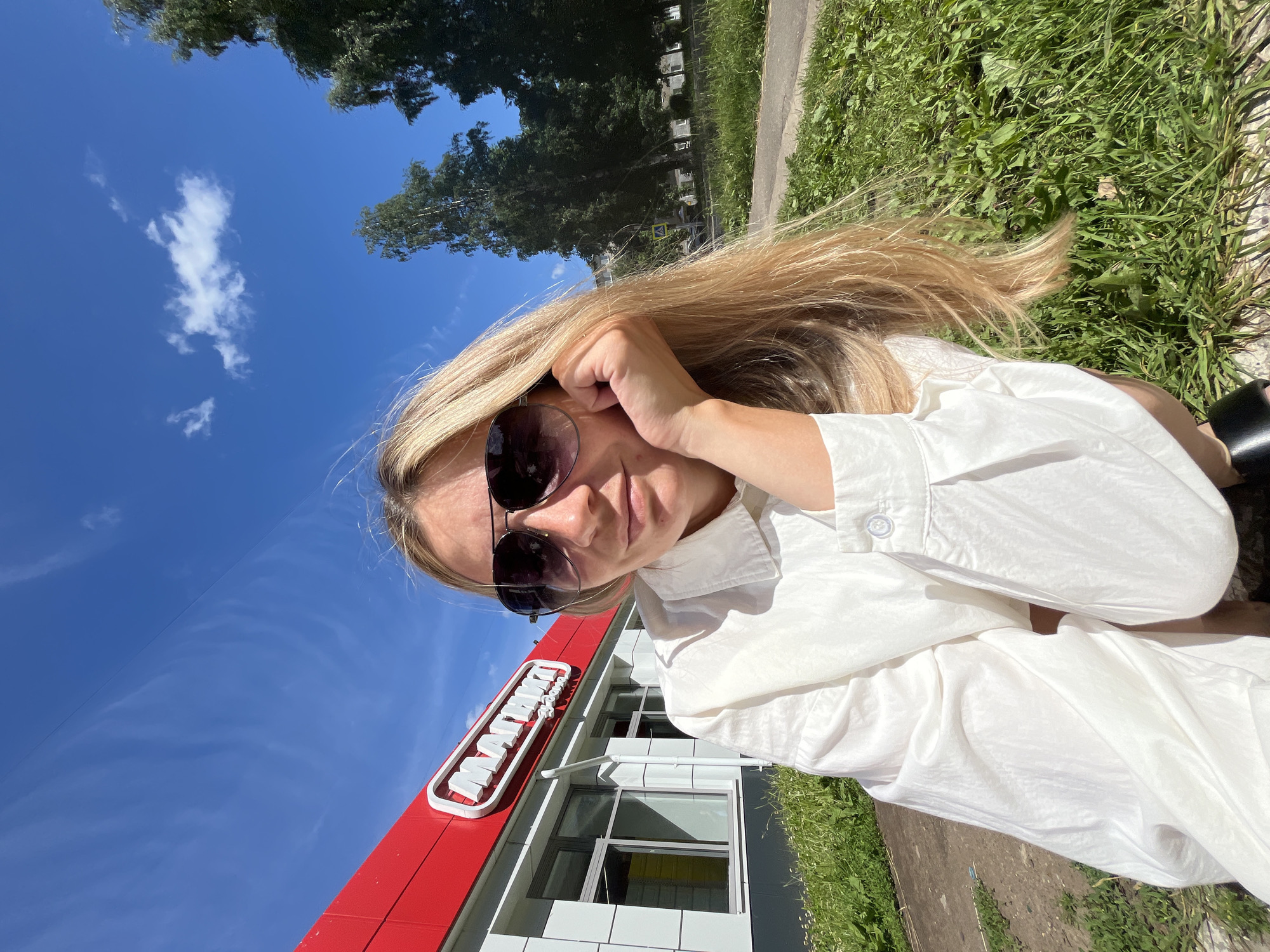 A fair-skinned woman with long blonde hair, wearing sunglasses and a white button-down shirt, sits in front of a "Magnit" store. The store has a red facade and white lettering. The woman is resting her chin on her hand and her hair is falling over her shoulder. She is positioned in the foreground, with the store and trees in the background. The image was taken on a sunny day, with a blue sky and scattered clouds. The location is the city of Chaikovsky, Russia.