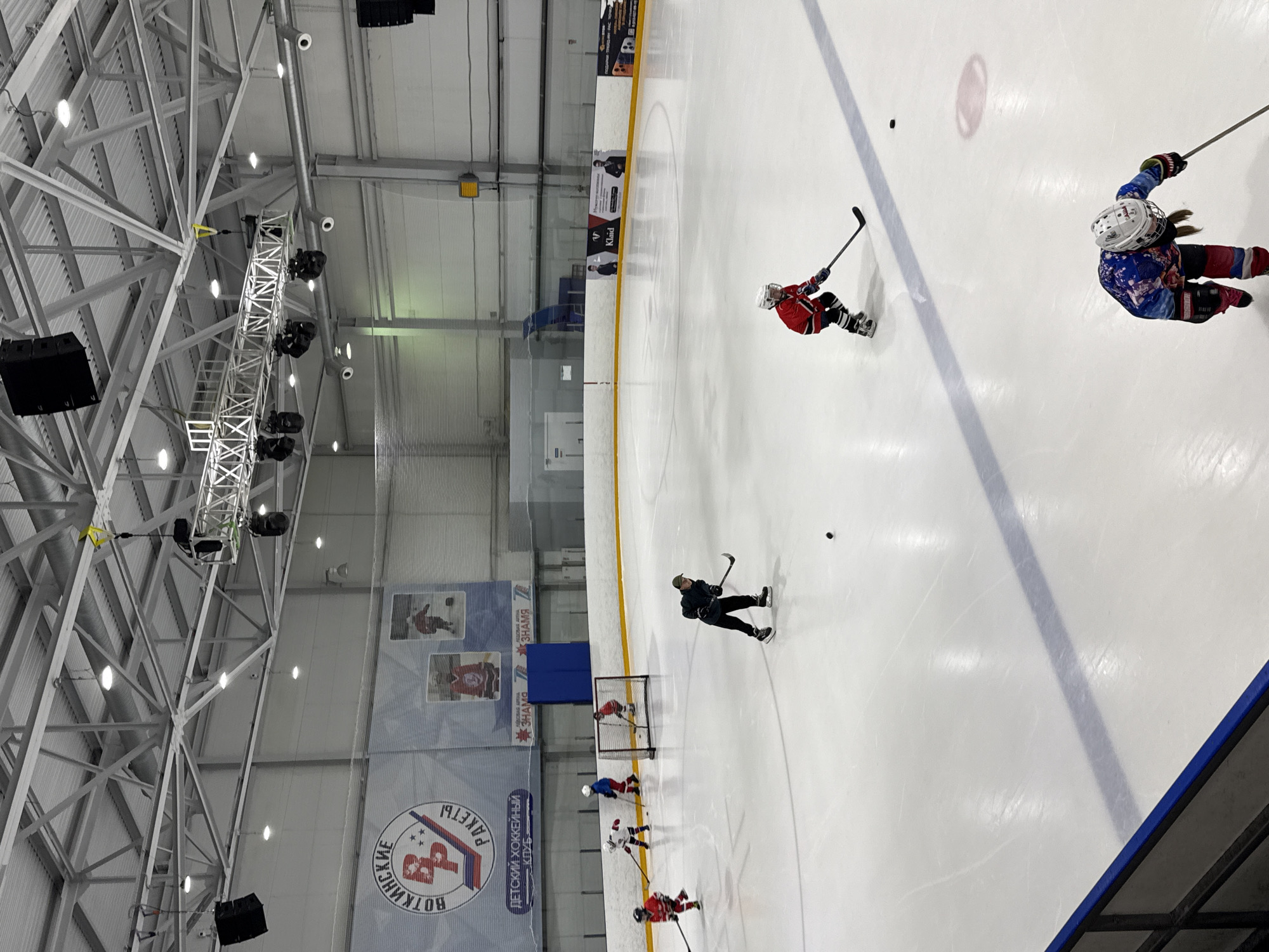 This is an aerial view of an indoor ice hockey rink in Votkinsk, Russia, likely part of a youth hockey club. The rink is brightly lit with overhead lights. On the ice, several young players in full hockey gear are practicing. One player in a red jersey is positioned near a net, another in a red and blue jersey is skating towards the center of the rink, and a player in a dark jacket and green cap is on the right side of the frame, facing left and appearing to be in motion. Another player in a red jersey with white and blue accents is skating from the top left of the frame. In the foreground, a player in a colorful jersey with a white helmet is skating towards the bottom right of the frame. There is a hockey puck visible on the ice. In the background, banners with the logo of "Votkinskiye Rakety" (Votkinsk Rockets) and other graphics are displayed along the rink's wall. The ceiling of the arena is a complex structure of metal beams and lighting fixtures.