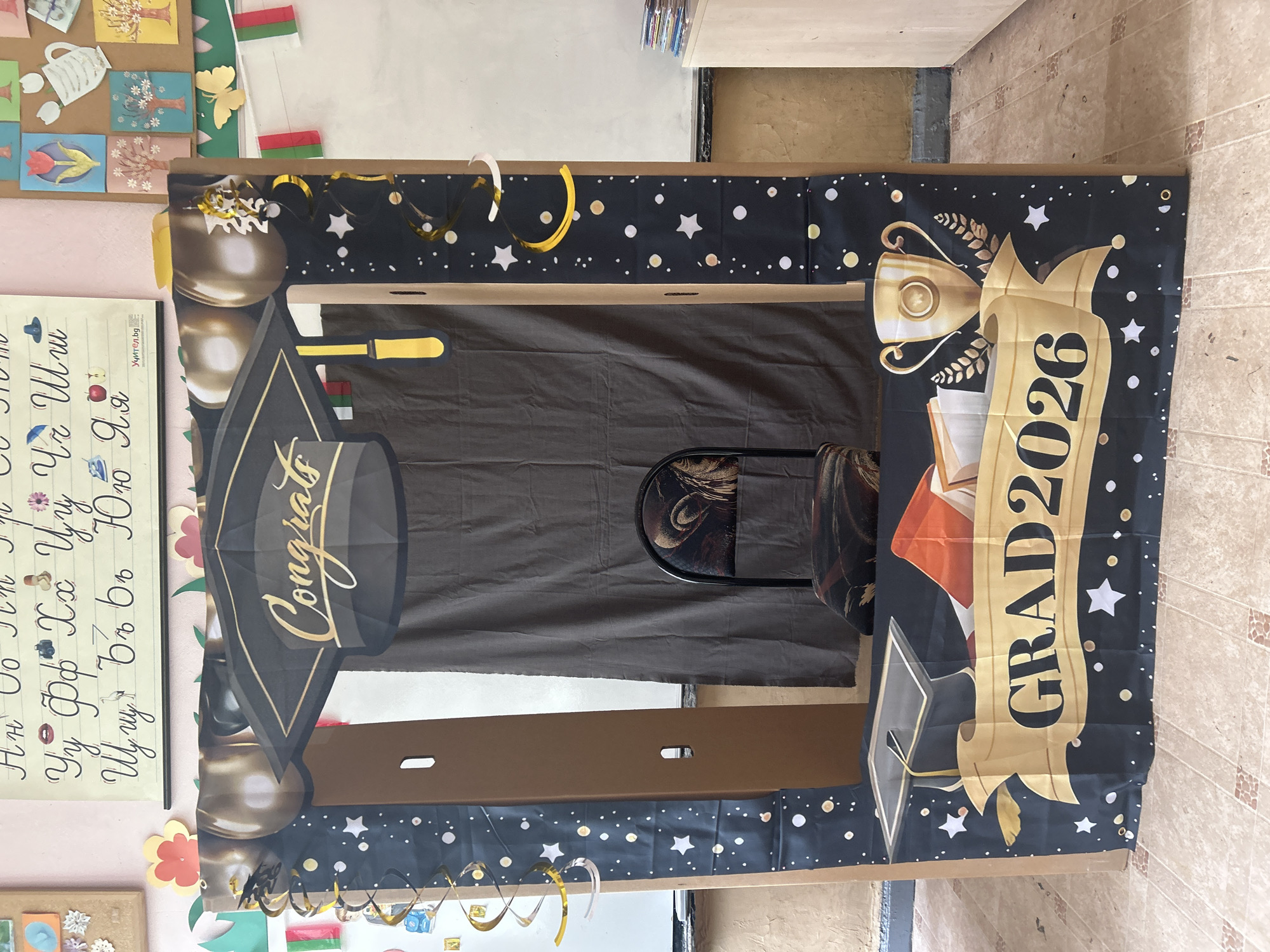The image shows a graduation photo booth made of cardboard, decorated with a black backdrop featuring gold stars, graduation caps, a trophy, and the text "GRAD 2026." The booth is set up indoors with a chair visible through the opening. Various educational charts and decorative items are visible on the wall to the left. The specific location is unclear from the image alone; there is no visible indicator of being in Obretenik, Bulgaria.