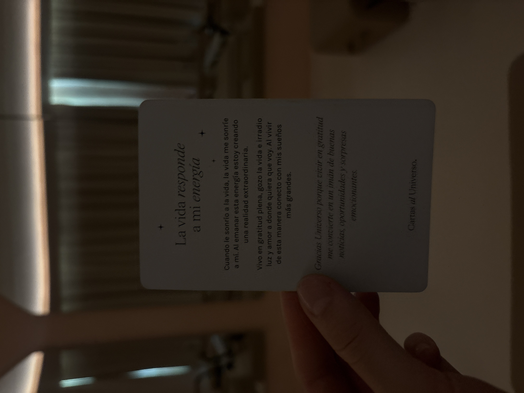 Here is a detailed description of the image:
**Scene:** A person's hand is holding up a card indoors in a dimly lit environment. The card is a rectangular shape with rounded corners, featuring text in Spanish. The background is blurred but appears to be a room with some light filtering through a window or opening.
**Card Details:** The card has text in black ink, arranged in multiple lines. The main title is "La vida responde a mi energía." The text below the title describes a positive affirmation or message related to energy, gratitude, and attracting good things in life. The card is identified as "Cartas al Universo."
**Hand:** The hand holding the card appears to be light-skinned. The fingers are partially visible, and the hand seems to be holding the card firmly but gently.
**Environment:** The environment is generally dark, but there is some light present, as the card and hand are partially illuminated. The background is out of focus, suggesting a shallow depth of field. The room appears to have some vertical structures.
**Location:** There is no explicit indication of the location within the image itself, let alone the city of Mexico City, Mexico. No identifying landmarks are visible.