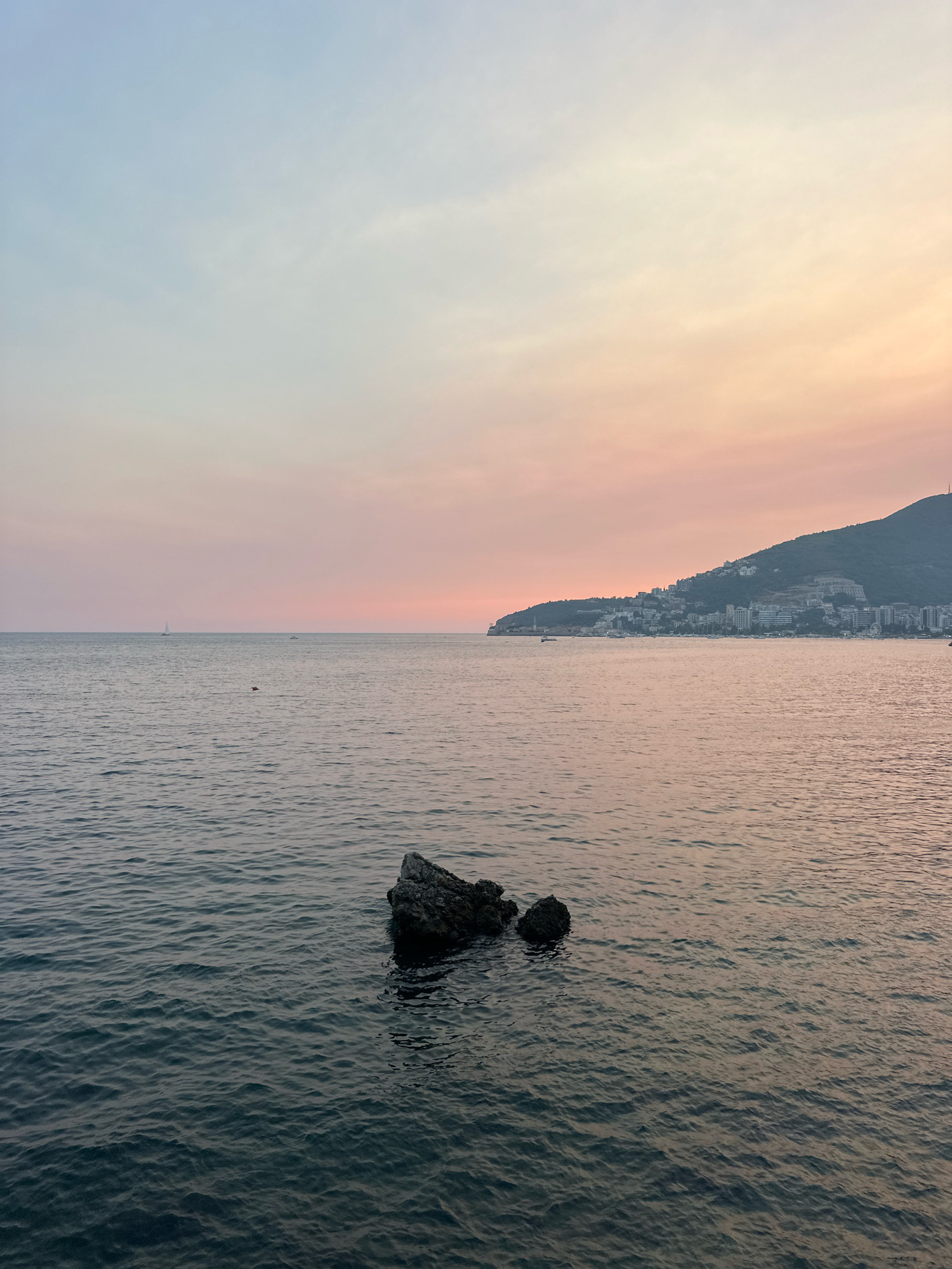 The image captures a serene seascape at dusk near the city of Budva, Montenegro. The sky is painted with soft hues of blue, pink, and orange, transitioning from light blue at the top to a warm, rosy glow near the horizon. A faint band of pink light is visible just above the distant shoreline.
The calm sea, with its dark, rippling surface, occupies the majority of the foreground. Two dark, weathered rocks protrude from the water in the middle ground, their surfaces wet and reflecting the muted light. In the distance, a white sailboat with its sail down is visible on the left side of the frame, its silhouette stark against the softer tones of the sea and sky. Further to the right, along the coast, is a cluster of buildings forming the town of Budva, nestled against the gentle slope of a green hill. The buildings appear mostly white and light-colored, with some darker structures visible as well. The coastline stretches across the frame, culminating in a dark, rounded hill at the far right. Several small boats are anchored or moving in the bay in front of the town. The overall impression is one of tranquility and natural beauty as the day ends.