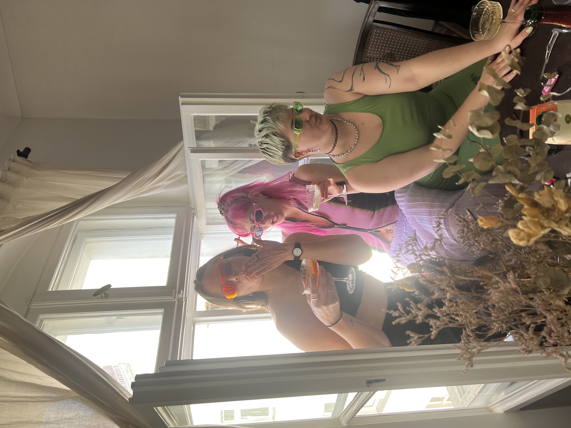 Three women are seated in a window frame. The woman on top is wearing a green tank top and green sunglasses. She has tattoos on her left arm and is holding a glass with a clear liquid. The woman in the middle has pink hair and is wearing pink sunglasses. She is holding a glass of champagne or sparkling wine. The woman on the bottom is wearing orange sunglasses and a black top. She is holding a glass with an orange liquid. There is a curtain hanging to the left of the window. There is a bouquet of dried flowers to the right. The location is unclear, but given the request of Berlin, Germany, one can deduce that the photo was taken indoors in a building in Berlin.