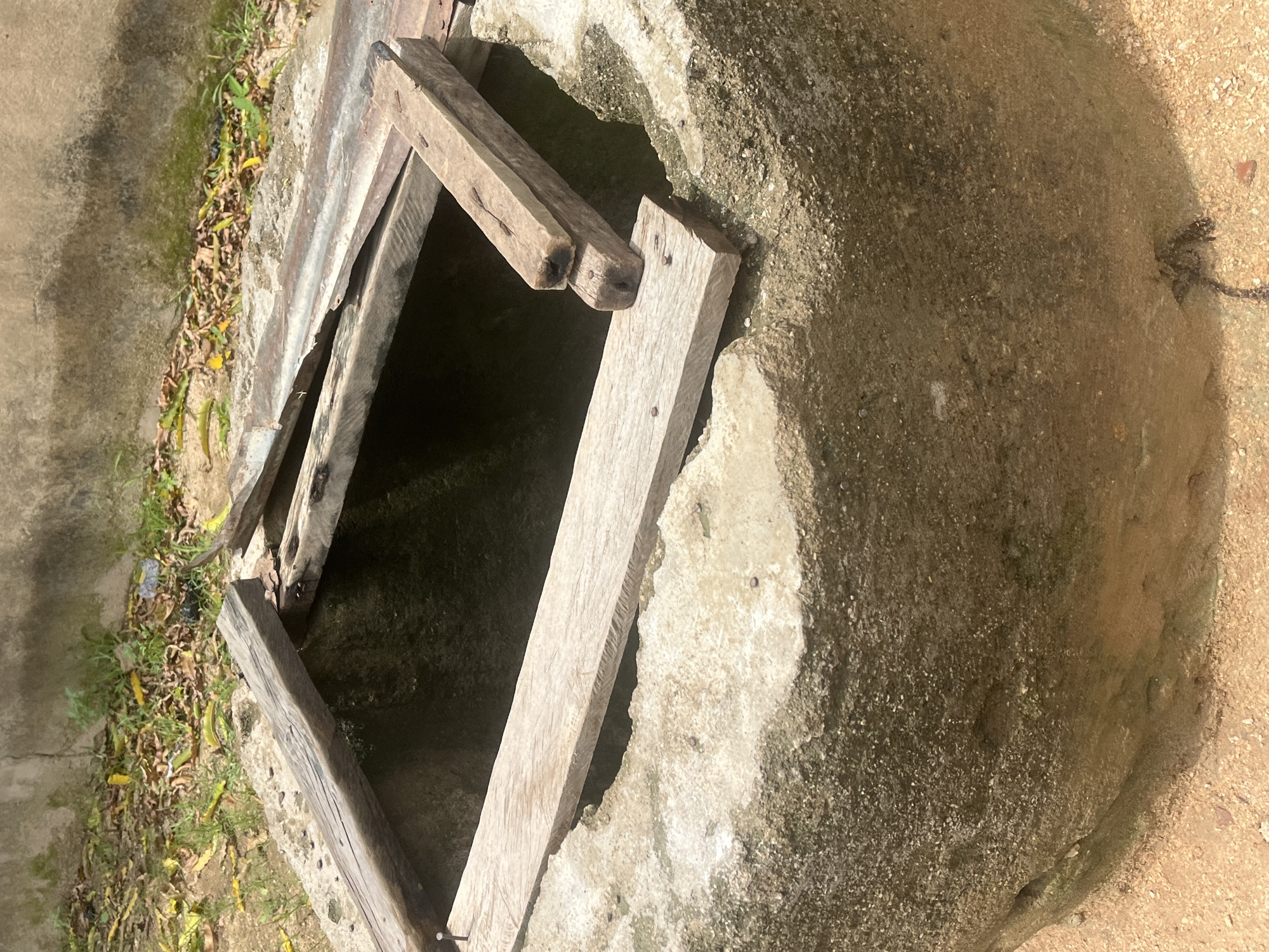 The image shows an old, circular well made of rough concrete. The well is open at the top, with several weathered wooden planks laid across it, some overlapping and seemingly unstable. The ground around the well is a mixture of sand and dirt, with some dry leaves and sparse green vegetation scattered nearby. A rough concrete wall is visible in the background, with patches of moss and discoloration. The lighting suggests it is daytime, with harsh shadows. The well appears to be located outdoors, in an area that may be somewhat neglected. This scene could be found in a rural or semi-urban setting in Jalingo, Nigeria, where such wells are often used as a source of water. There are no people or animals visible in the image.