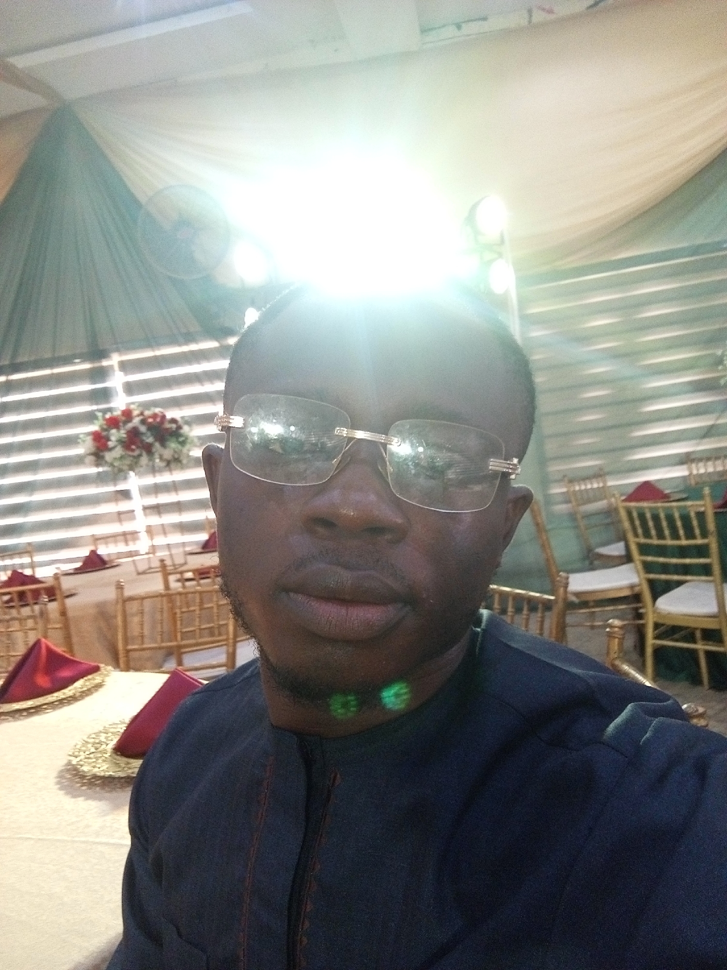 The image is a selfie taken by a dark-skinned man wearing glasses and a dark blue garment with red embroidery. He is seated at a table in what appears to be an event space, possibly a wedding reception or a banquet. The setting is decorated with draped fabric in neutral tones and accented with floral arrangements. Gold chairs are visible around tables covered with light-colored tablecloths, and some tables have burgundy napkins. The lighting is bright, with a strong glare suggesting either direct sunlight or powerful artificial lighting. The overall atmosphere seems festive and formal. The man's facial expression is neutral. There is no visible text in the image. The context provided indicates the location is Abuja, Nigeria.