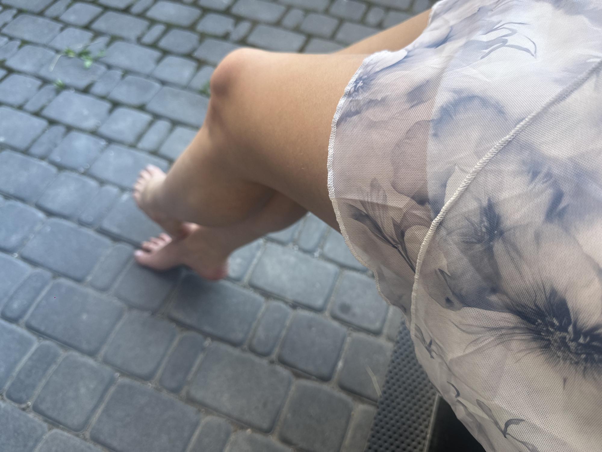A close-up, high-angle shot shows a person's legs and feet as they sit on a textured surface. The person is wearing a sheer, light-colored dress with a delicate floral pattern in shades of blue and grey. The fabric of the dress appears gauzy and slightly translucent, with visible stitching along the hem. One leg is bent at the knee, with the knee cap clearly visible. The other leg is extended slightly.
The person is barefoot, and their feet are resting on a grey paved surface composed of rectangular and square stones. The paving stones have a rough, textured appearance and are laid in a regular pattern. There are small patches of green vegetation growing between some of the stones, suggesting an outdoor location. The lighting suggests it is daytime, with diffused light creating soft shadows. The overall mood appears casual and relaxed. No text is visible in the image.