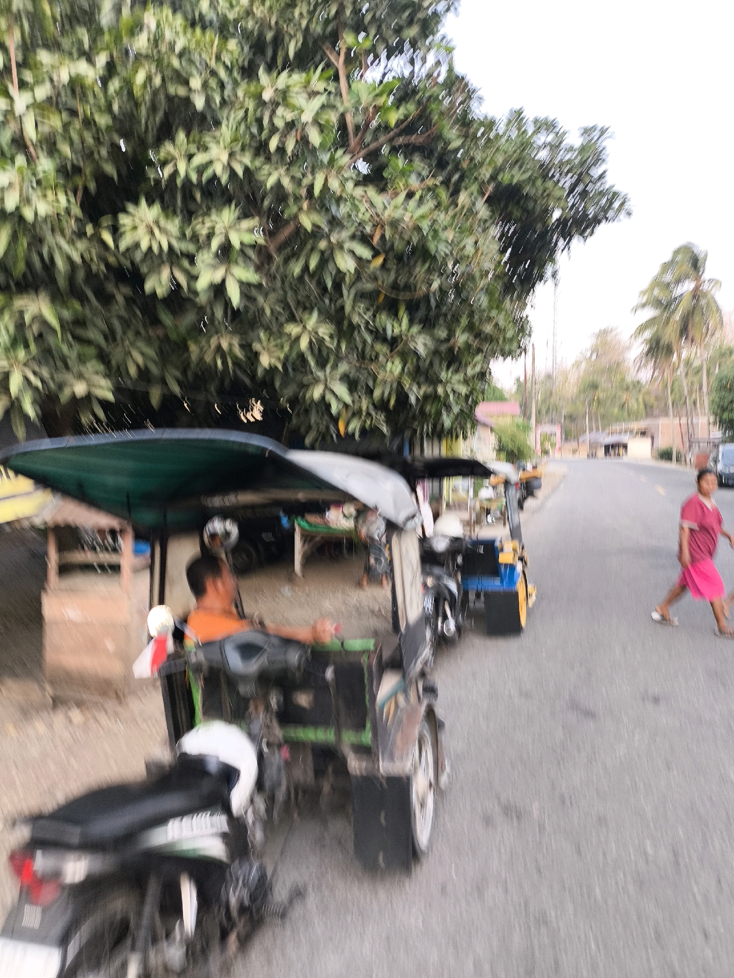 In the city of Unknown, Indonesia, a blurry, daytime scene unfolds along a paved road bordered by lush green foliage. In the foreground, a three-wheeled vehicle, possibly a becak, is parked. A person wearing an orange shirt sits in the driver's seat. To the left, a motorcycle is partially visible. Further down the road, another three-wheeled vehicle, this one with a yellow frame, is also parked. A woman wearing a pink shirt and shorts walks across the road from right to left. In the background, houses and palm trees are visible under a bright sky. The motion blur suggests the photograph was taken from a moving vehicle.