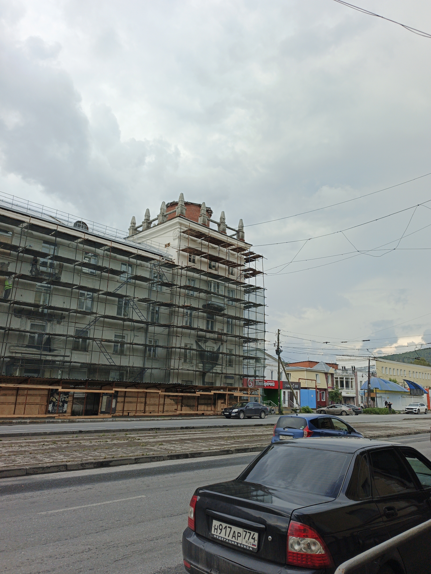 The image depicts a street scene in Zlatoust, Russia, featuring a multi-story building undergoing renovation, characterized by extensive scaffolding. The building is a prominent structure on the left side of the frame, its facade covered in scaffolding and protective netting. A single person wearing a yellow vest is visible on the scaffolding. Below the building, there's a wooden structure, possibly a temporary market or barrier.
The street in the foreground is paved, with visible tram tracks running parallel to the sidewalk. Several cars are present: a black sedan in the immediate foreground, a blue hatchback further down the street, and a dark gray sedan also in traffic. In the background, a row of shops and smaller buildings lines the opposite side of the street, some with awnings and signs, including one that reads "Пятёрочка" (Pyaterochka), a Russian supermarket chain. Power lines and utility poles are visible overhead, contributing to the urban environment. The sky is overcast with dense clouds, suggesting a cloudy or potentially rainy day. The overall atmosphere is that of an active urban area with ongoing construction.