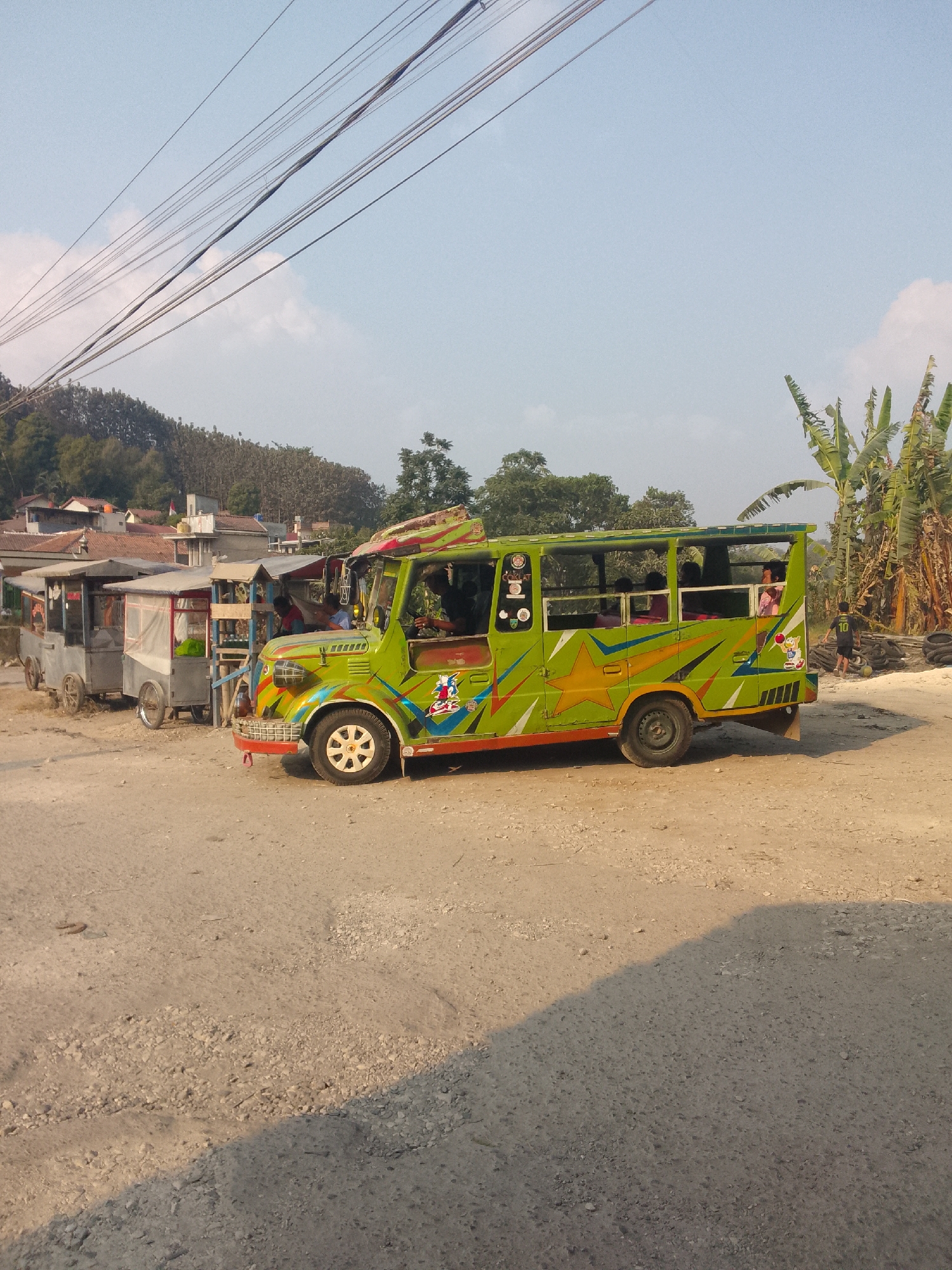 In Ciburuy, Indonesia, a brightly decorated green bus with cartoon decals is parked on a dirt road. The bus has an open-air design with passengers visible inside. To the left of the bus are three food stalls on wheels, also parked on the dirt road. In the background, there are houses nestled on a hillside covered with trees. The sky is clear with a few wispy clouds. A young boy wearing a black shirt with the number 10 is standing near a pile of tires on the right side of the image. Overhead, power lines stretch across the frame.