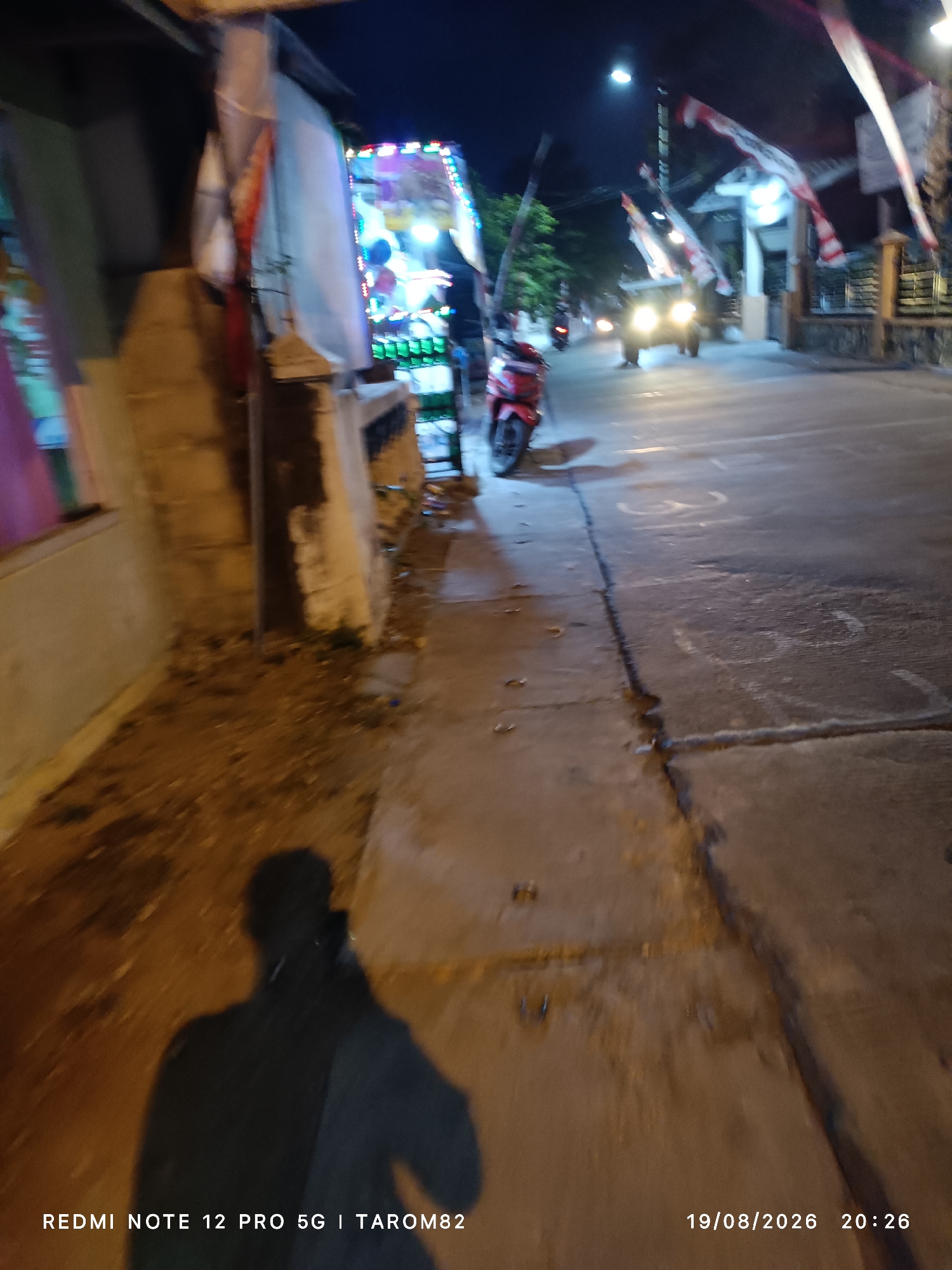 The image shows a street scene at night in Sidodadi, Indonesia. The foreground is dominated by a shadow of a person, possibly the photographer, cast on the ground. To the left, there is a section of a building with a wall and an open window or doorway. Decorations, possibly for a celebration, including balloons and string lights, are visible on the side of the building. In the mid-ground, a red motorcycle is parked on the side of the road. Further down the street, a vehicle, likely a car or small truck, is visible with its headlights on. Several Indonesian flags are seen hanging above the street, suggesting a national holiday or event. The road surface is asphalt, and there are painted markings on it. The overall atmosphere is dimly lit, typical of a nighttime urban environment. The date and time stamp indicate the photo was taken on August 19, 2026, at 20:26.