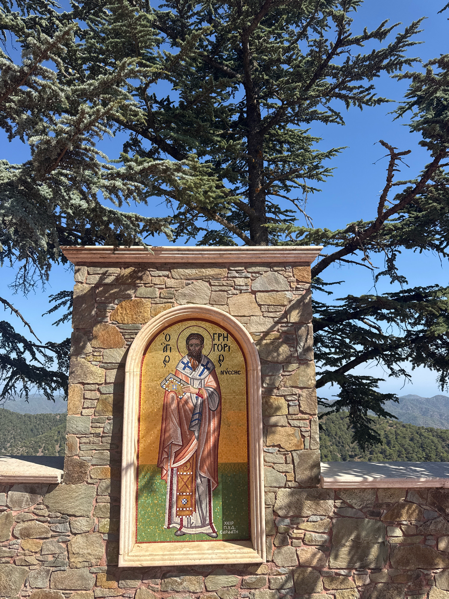 The image captures a mosaic of a religious figure set within an arched stone structure. The mosaic depicts a man with a beard, wearing religious vestments and holding a book. Greek text is visible above and below the figure. The stone structure is made of rough-hewn rocks of various colors, including brown, tan, and gray. This structure is situated outdoors, under the branches of a large evergreen tree with dark green needles. In the background, a clear blue sky transitions to a landscape of rolling hills covered in green vegetation, suggesting a mountainous or hilly terrain. The setting appears to be in a rural or natural environment, consistent with the location of Tsakistra, Cyprus, which is known for its mountainous landscape and natural beauty. The lighting suggests a bright, sunny day, casting shadows on the stone structure.