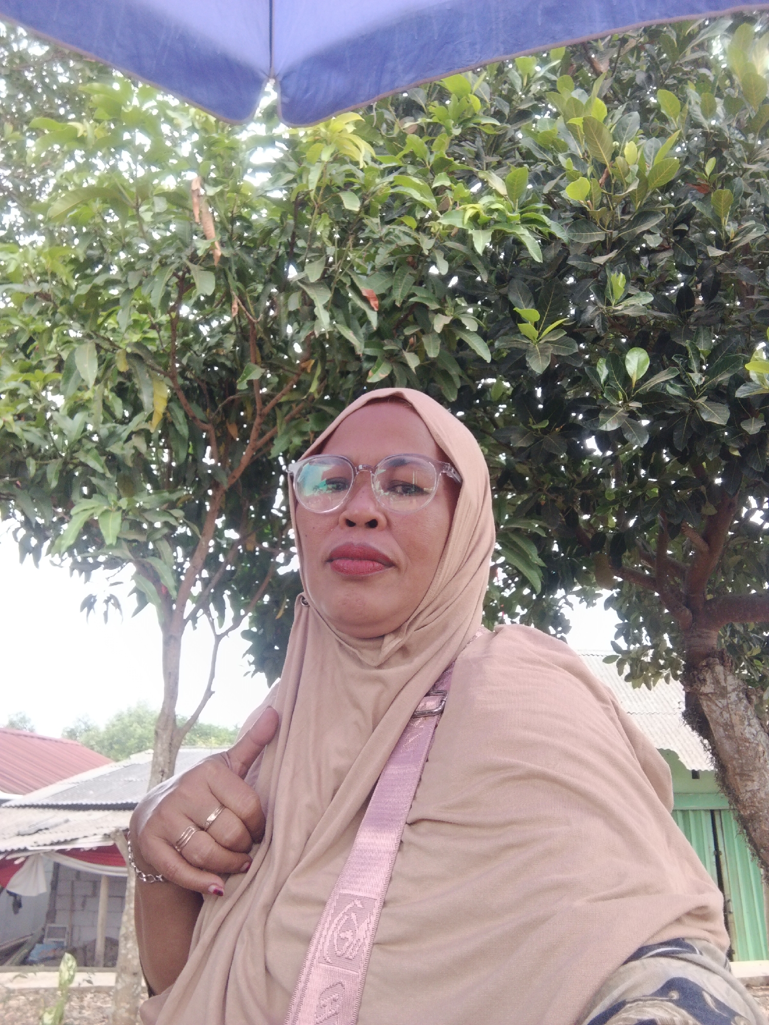 A woman is taking a selfie outdoors in Warungkiara, Indonesia. She is wearing a light brown hijab and glasses, with a pink strap from her bag visible over her shoulder. Her right hand is raised, showing rings on her fingers, and she is giving a thumbs up. In the background, there is lush green foliage from trees and a glimpse of buildings with corrugated metal roofs. The overall impression is a casual, outdoor snapshot.