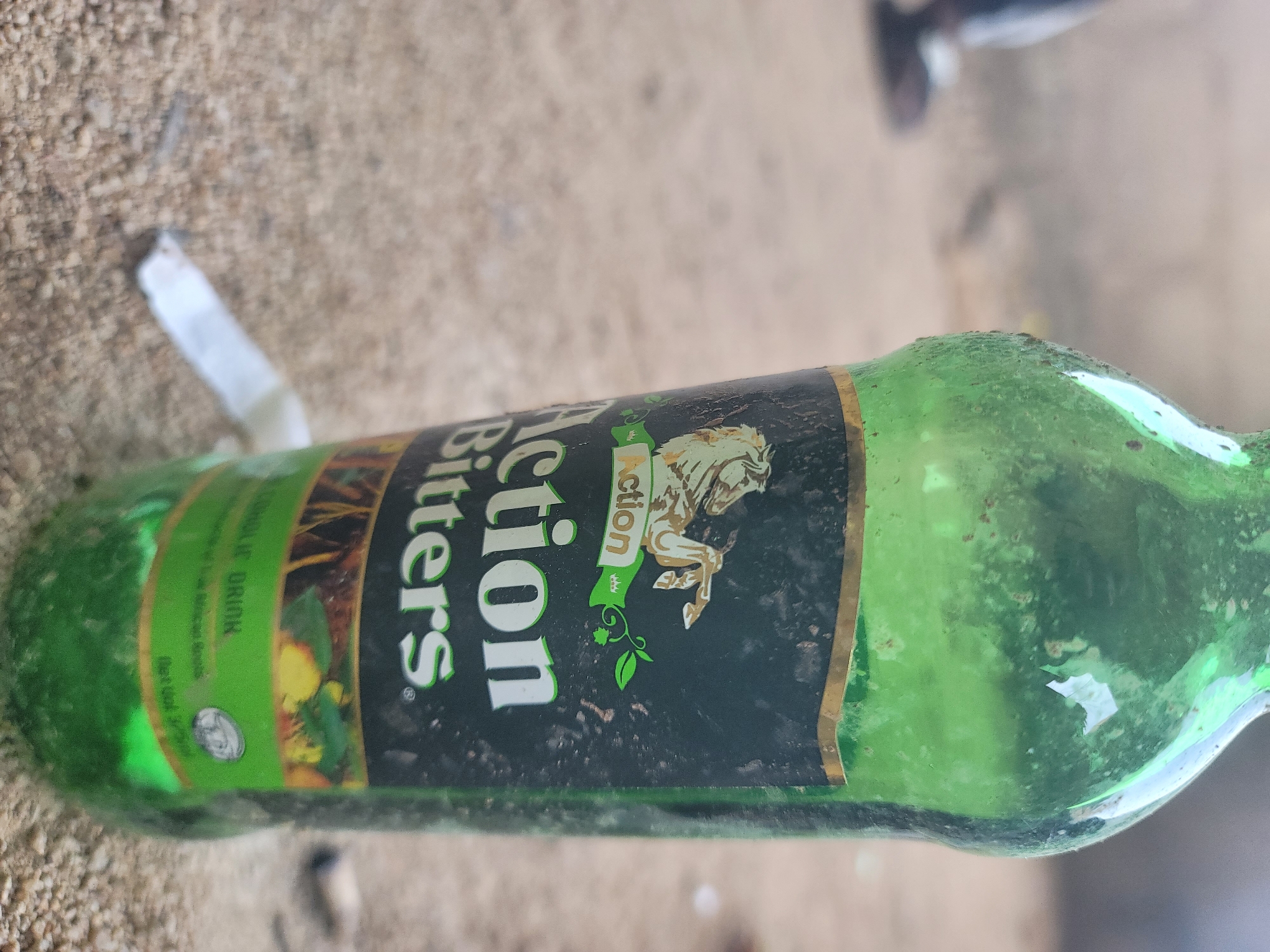 The image shows a close-up of a green glass bottle of "Action Bitters" alcoholic drink. The bottle is partially obscured by the camera's focus and is resting on a sandy surface, possibly in an outdoor setting. A partially smoked cigarette butt is visible in the foreground, near the base of the bottle. In the blurred background, a person's legs and feet are visible, wearing dark trousers and sandals. The overall scene appears to be a casual or possibly unkempt outdoor environment.