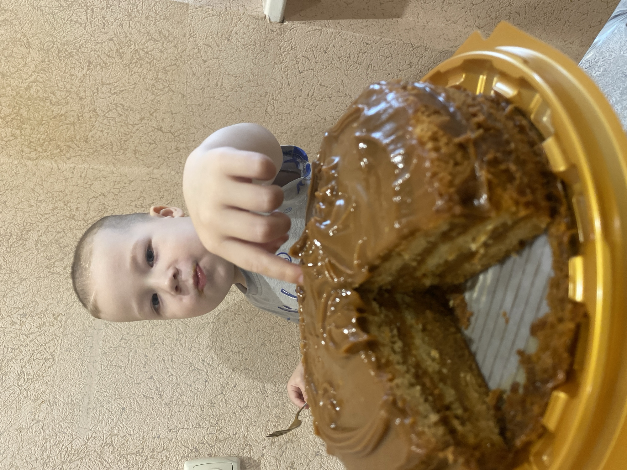 A young child is reaching for a piece of caramel-covered cake in a yellow plastic container. The child is fair-skinned with short hair and is wearing a gray shirt. The background is a light beige textured wall. A fork is in the child's other hand. There is no information in the image to suggest the location is Sochi, Russia.