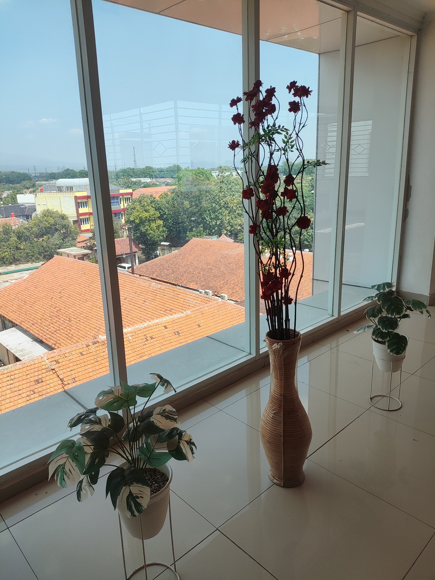 The image depicts an interior scene with a large window overlooking a cityscape. Inside, there are two potted plants and a tall, woven vase containing artificial red flowers. The plant on the left has large, variegated leaves of green and white. The plant on the right is smaller and appears to be a similar variety. The vase is light brown and has a textured, vertical pattern. The floor is tiled in a beige color, with visible grout lines.
Through the window, a view of Cirebon, Indonesia, is visible. The cityscape includes buildings with orange tile roofs, some taller structures with yellow and red facades, and lush green trees. In the distance, hazy hills or mountains can be seen, along with a clear blue sky. The large window features vertical mullions that divide the view. The overall impression is one of an indoor space with a view of an urban environment in Cirebon.