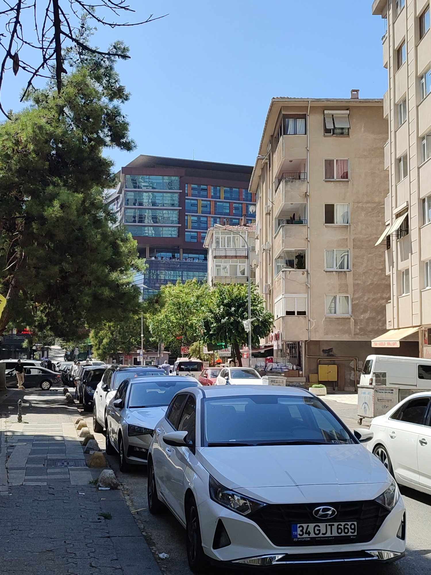 This is a street scene in Kadıköy, Turkey, on a sunny day. The foreground is dominated by a white Hyundai car parked on the right side of the street, facing forward. The car has a license plate reading "34 GJT 669". To its right, another white car is partially visible, also parked. The street is lined with parked cars, creating a sense of urban density.
Behind the parked cars, there are buildings of varying architectural styles. To the right, a light-colored apartment building with balconies and numerous windows stands tall. Further back, a modern building with a glass facade, featuring colored panels in shades of blue, orange, and brown, is visible, reflecting the sky and surroundings. Trees, including a large pine tree on the left, line the street, casting shadows on the sidewalk.
The sidewalk is paved with stone tiles and concrete. A few people are visible walking on the sidewalk and in the street, seemingly going about their daily activities. A white van is parked further down the street. The sky is clear and blue. The overall atmosphere is that of a typical urban environment in a Turkish city, with a mix of older and newer buildings and a steady flow of everyday life.