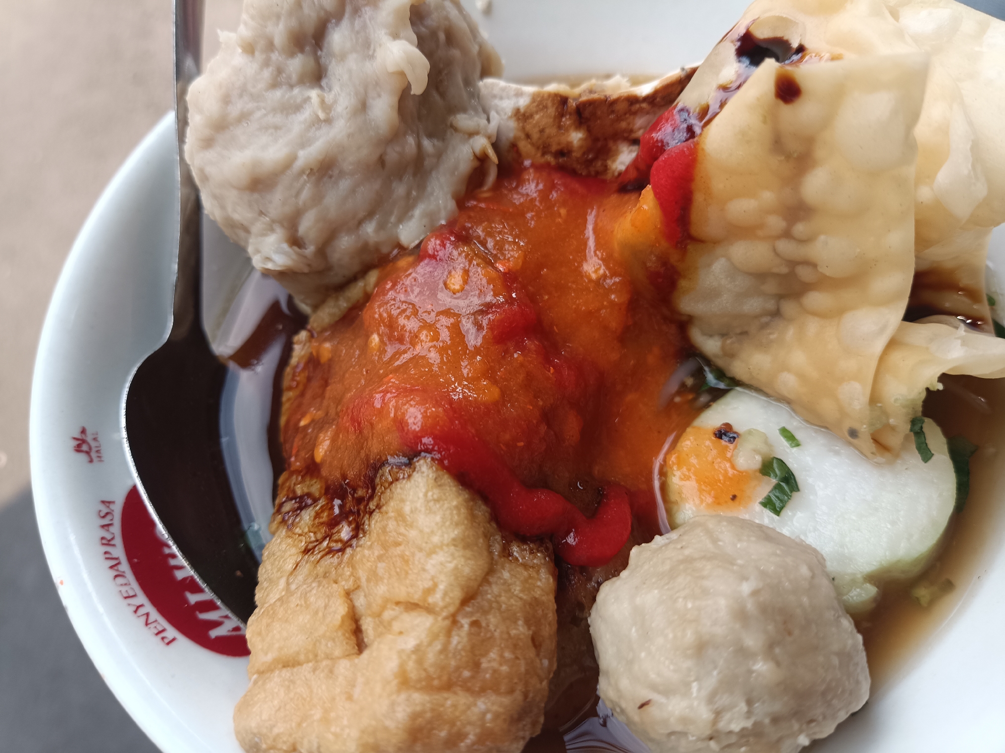 A close-up, overhead shot shows a bowl of Indonesian meatballs, or 'bakso', in a savory broth. The bowl is white with red lettering that reads "PENYEDAP RASA" and a "HALAL" symbol, suggesting it is from a restaurant in Jember, Indonesia. The bakso are light brown and spherical, some submerged in the dark broth, others peeking out. A dollop of vibrant red chili sauce is slathered over some of the meatballs, with thin streams of dark brown sauce drizzled on top. Crispy, golden-brown fried tofu cubes are visible, along with a sliced hard-boiled egg with a bright orange yolk. A shiny silver spoon rests on the left edge of the bowl, partially submerged in the broth. The background is out of focus, suggesting an outdoor or casually lit setting.