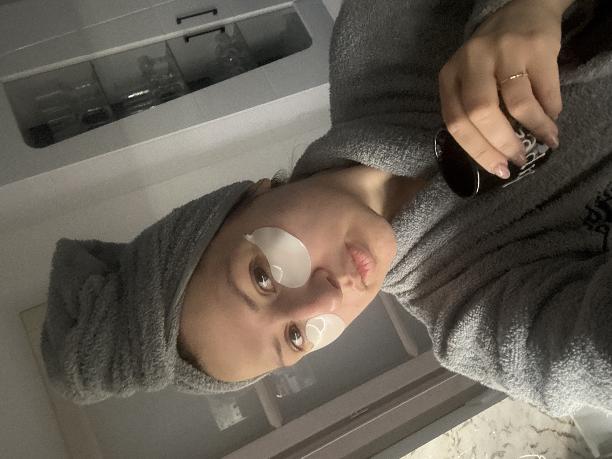 A woman in a gray robe and hair towel is holding a black mug. She has white under-eye patches on her face. In the background is a white cabinet with clear glass compartments, containing various glassware. The specific location in the city of Sofia, Bulgaria, cannot be determined from the visual information in the image.