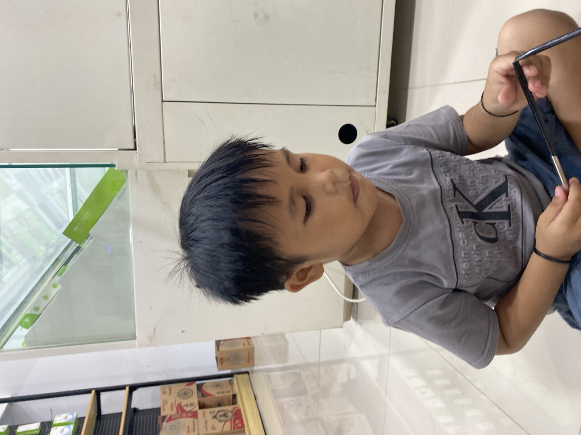 In Ta', Indonesia, a young boy sits on a tiled floor in what appears to be a retail environment. He is wearing a grey t-shirt with "Klein Calvin" text and a large "K" logo, along with blue shorts. The boy is looking down and holding a black object, possibly a frame or a thin rod, in his hands. In the background, there is a white cabinet with two doors, a glass display case with green accents, and shelves with boxes. The lighting suggests an indoor setting with artificial lights.