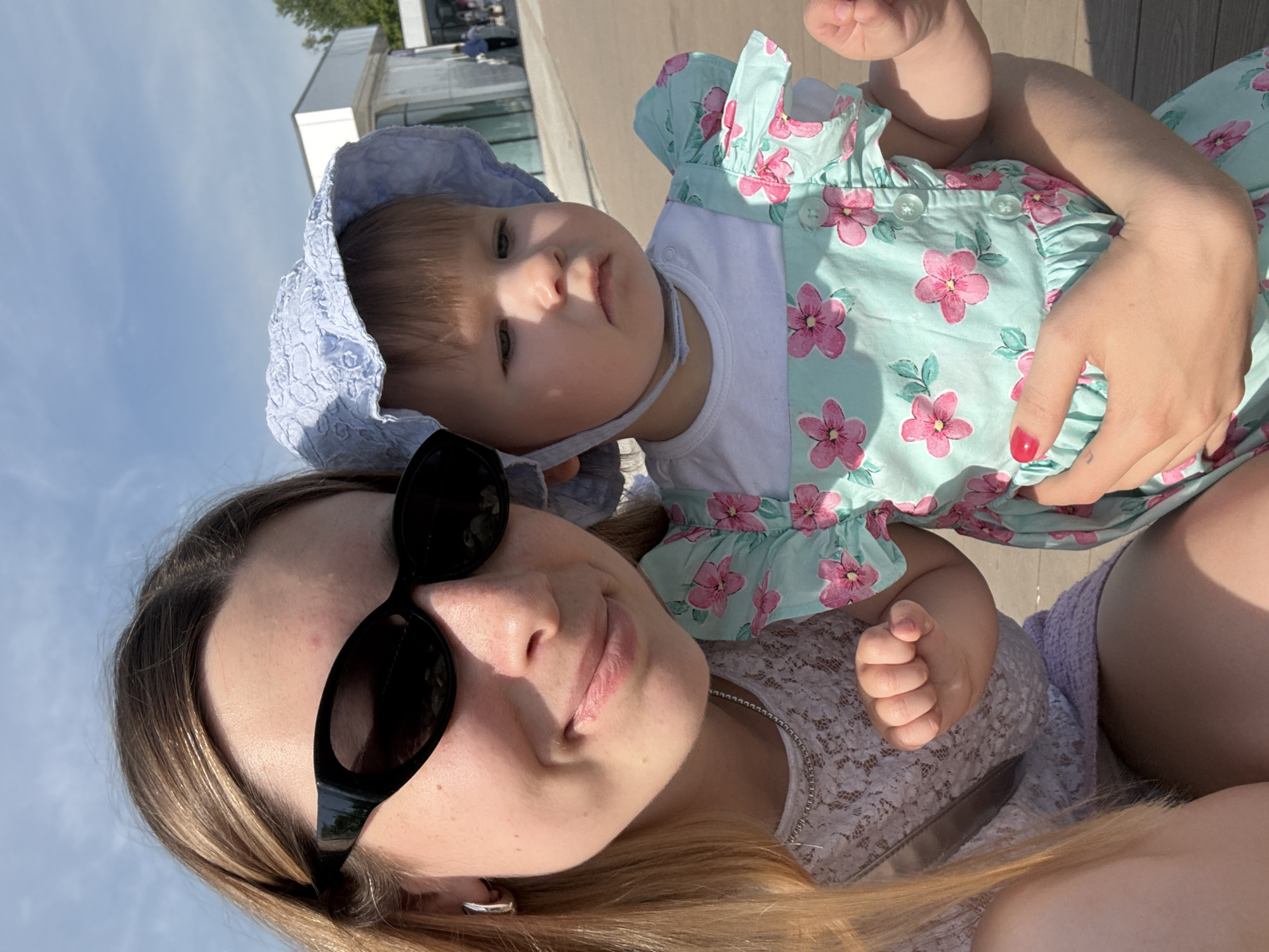 An outdoor, sunlit selfie captures a young woman holding a baby. The woman, positioned on the left side of the frame, has light brown hair parted in the middle and wears black cat-eye sunglasses. She is wearing a light-colored, possibly white, lace top and a thin silver chain necklace. A silver hoop earring is visible in her left ear. She exhibits a faint smile. Her left hand, with nails painted red, supports the baby.
The baby, held by the woman, is on the right. The baby has light brown hair and wears a light blue, textured bonnet-style hat secured with a chin strap. The baby is dressed in a white t-shirt layered under a light blue sundress adorned with a pink floral pattern and small light blue buttons down the front. The baby has a neutral, slightly pensive expression, looking towards the right of the frame.
The background shows a clear blue sky with minimal clouds at the top. The ground beneath them is a light brown, flat, paved surface, resembling a boardwalk or promenade. In the mid-ground, several modern, minimalist buildings with light-colored facades and large glass panels are visible. People can be seen in the distance near these structures, and green trees are present in the far background. The contemporary architectural style of the buildings and the design of the public space are characteristic of renovated recreational areas in Moscow. Based on these details, the scene is likely situated in Gorky Park in Moscow, Russia.