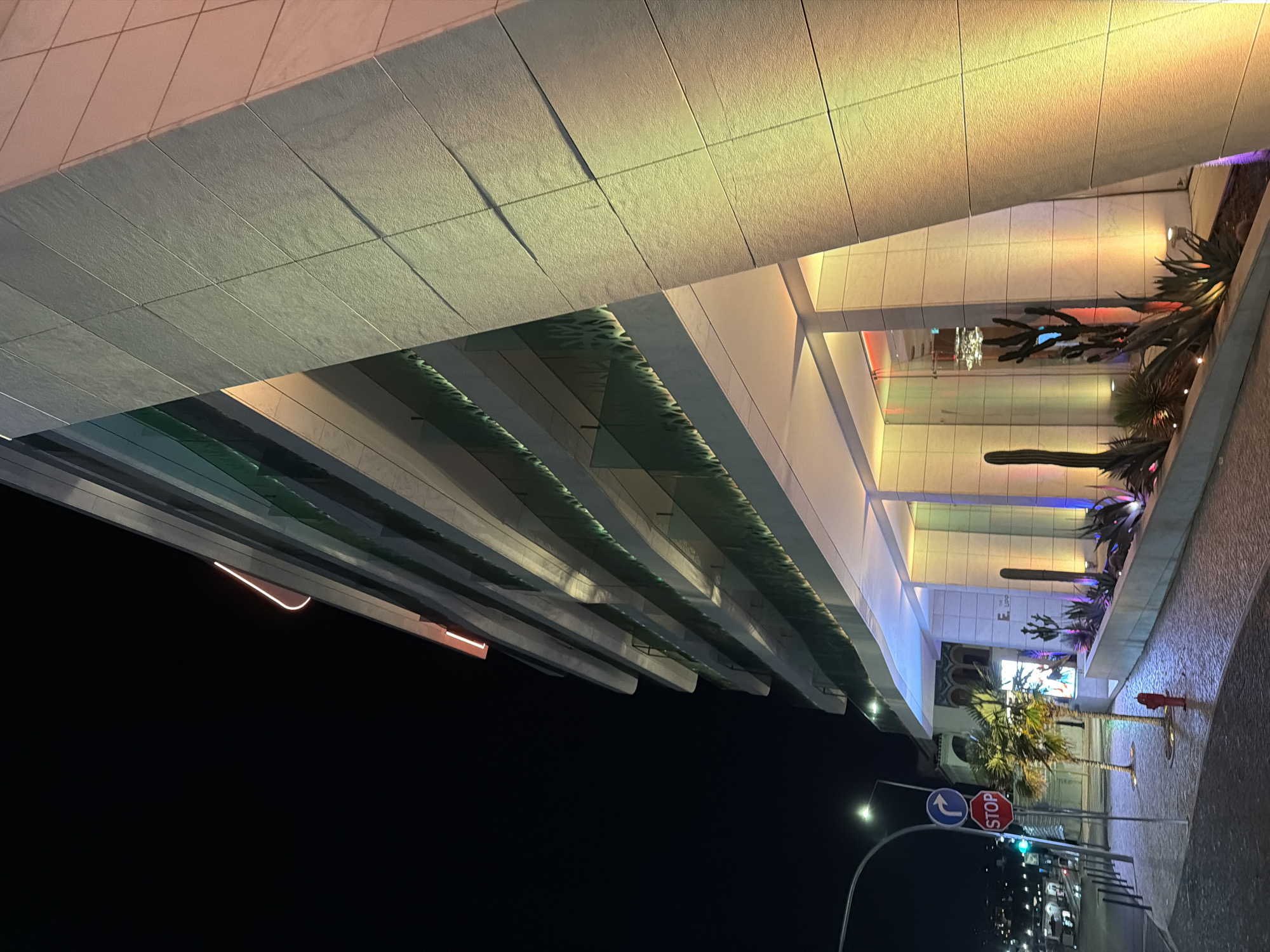 The image depicts the exterior of a modern, multi-story building at night in Cascais, Portugal. The building features a light-colored, textured facade with horizontal banding. It has multiple levels with glass balconies, and some levels appear to have decorative, green foliage or screening. The building is illuminated with colorful lights, predominantly yellow, orange, and pink on the facade, and blue and purple lights at the base, highlighting planters with various succulents and cacti.
In the foreground, a paved sidewalk and street are visible. A traffic signal with a red "STOP" sign and a blue directional arrow is mounted on a pole on the left. A red fire hydrant is located near the sidewalk's edge. Two small palm trees are planted near the building's entrance. In the background, streetlights and other buildings are faintly visible, suggesting an urban setting. The sky is dark, indicating it is nighttime. The overall scene is a well-lit, contemporary architectural structure in a coastal town.