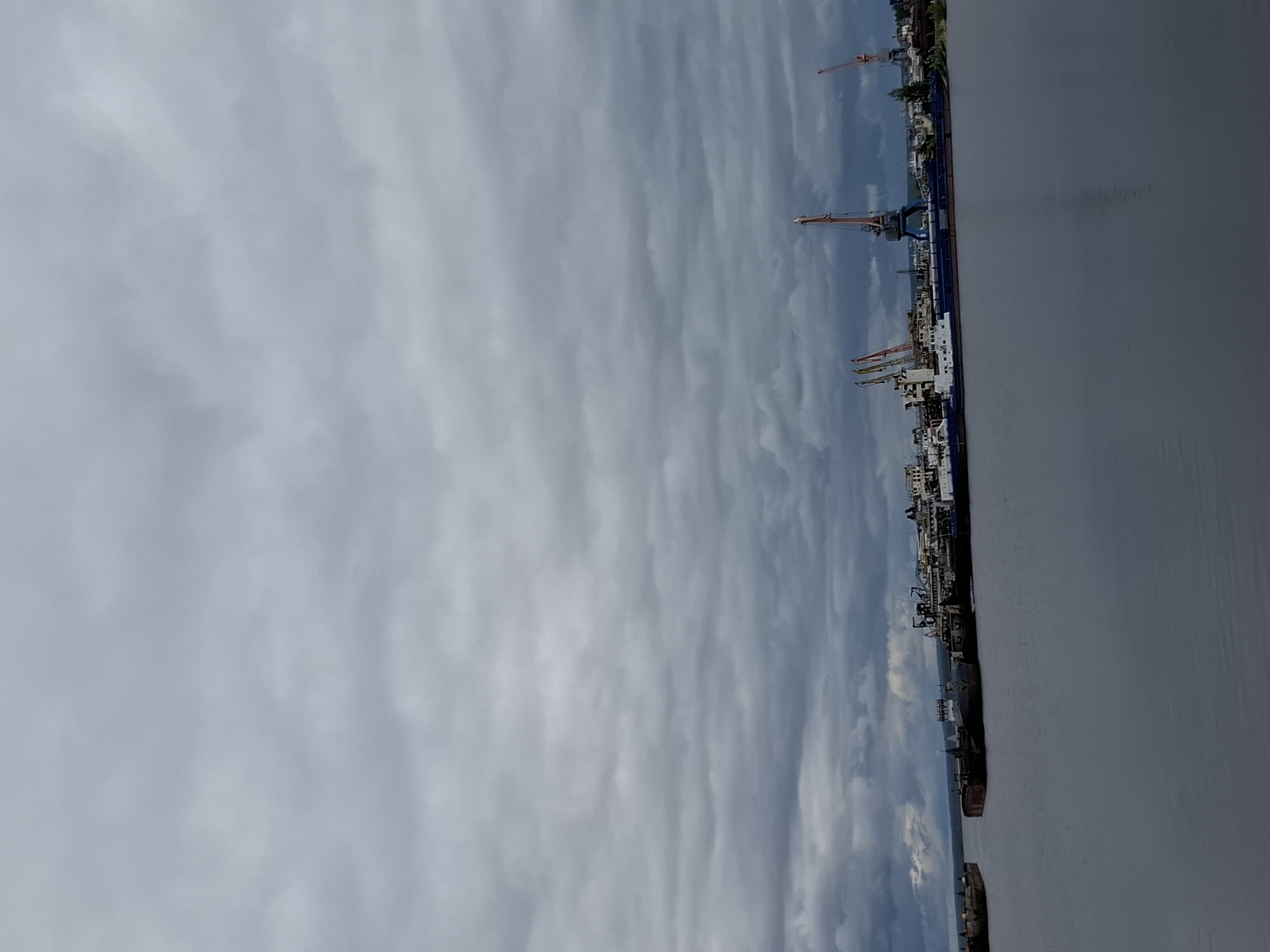The image shows a view of the city of Chaikovsky, Russia, on the banks of the Kama River. The sky is overcast with layered clouds. Along the riverbank are cranes and industrial structures, indicating a port or industrial area. The river itself occupies the lower portion of the image, appearing gray and calm. There are no visible people.