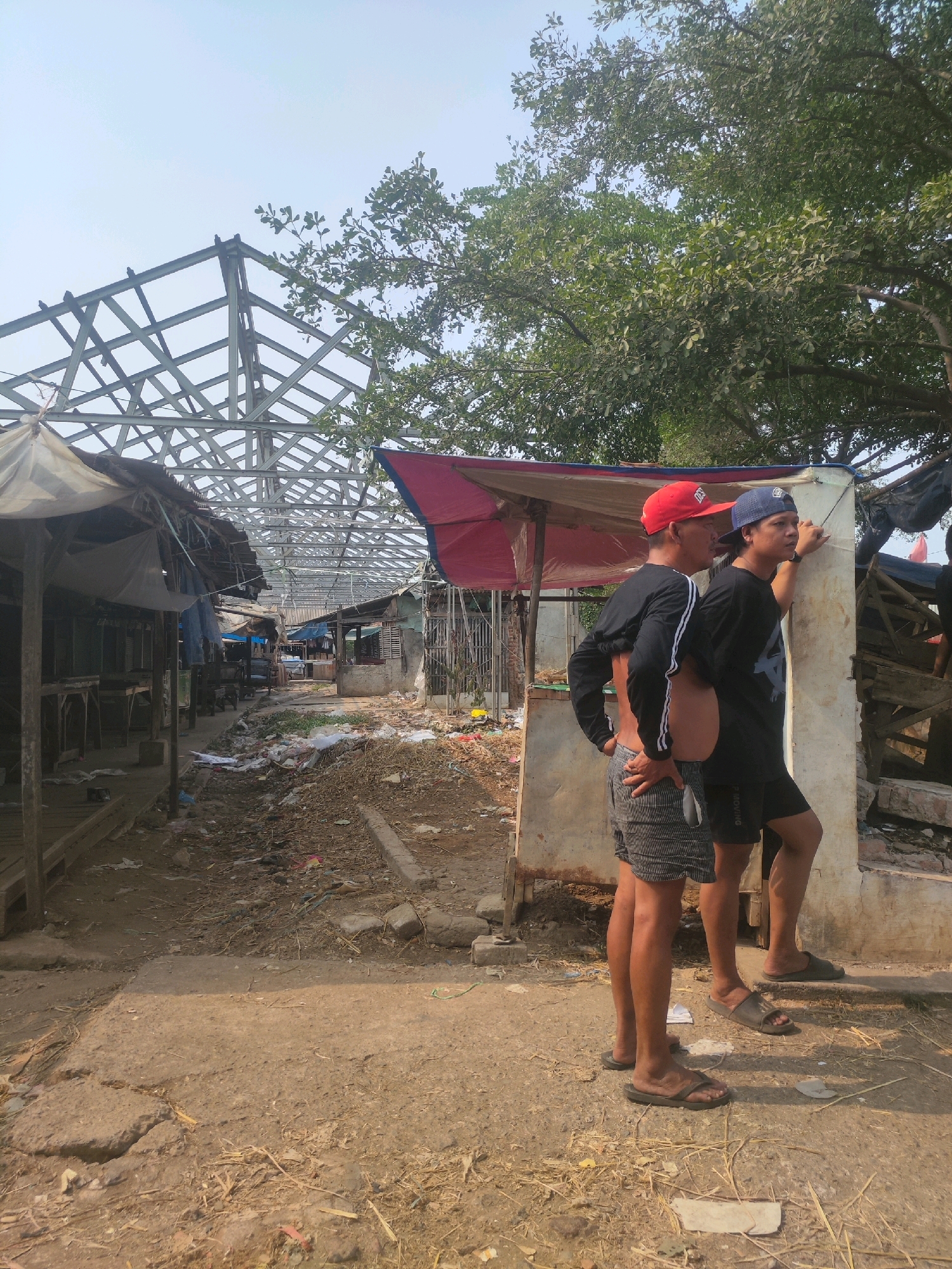 Two men stand facing a rundown market area in Karawang, Indonesia. The market appears to be under construction or renovation, with exposed metal framing for a roof visible above dilapidated stalls. The ground is dirt and debris-strewn, with scattered trash and building materials. One man, wearing a red baseball cap and a black long-sleeved shirt, stands with his hands on his hips. The other man, wearing a blue baseball cap and a black t-shirt, leans against a support beam. Both men are wearing shorts and sandals. The sky is clear and bright, suggesting a daytime scene.