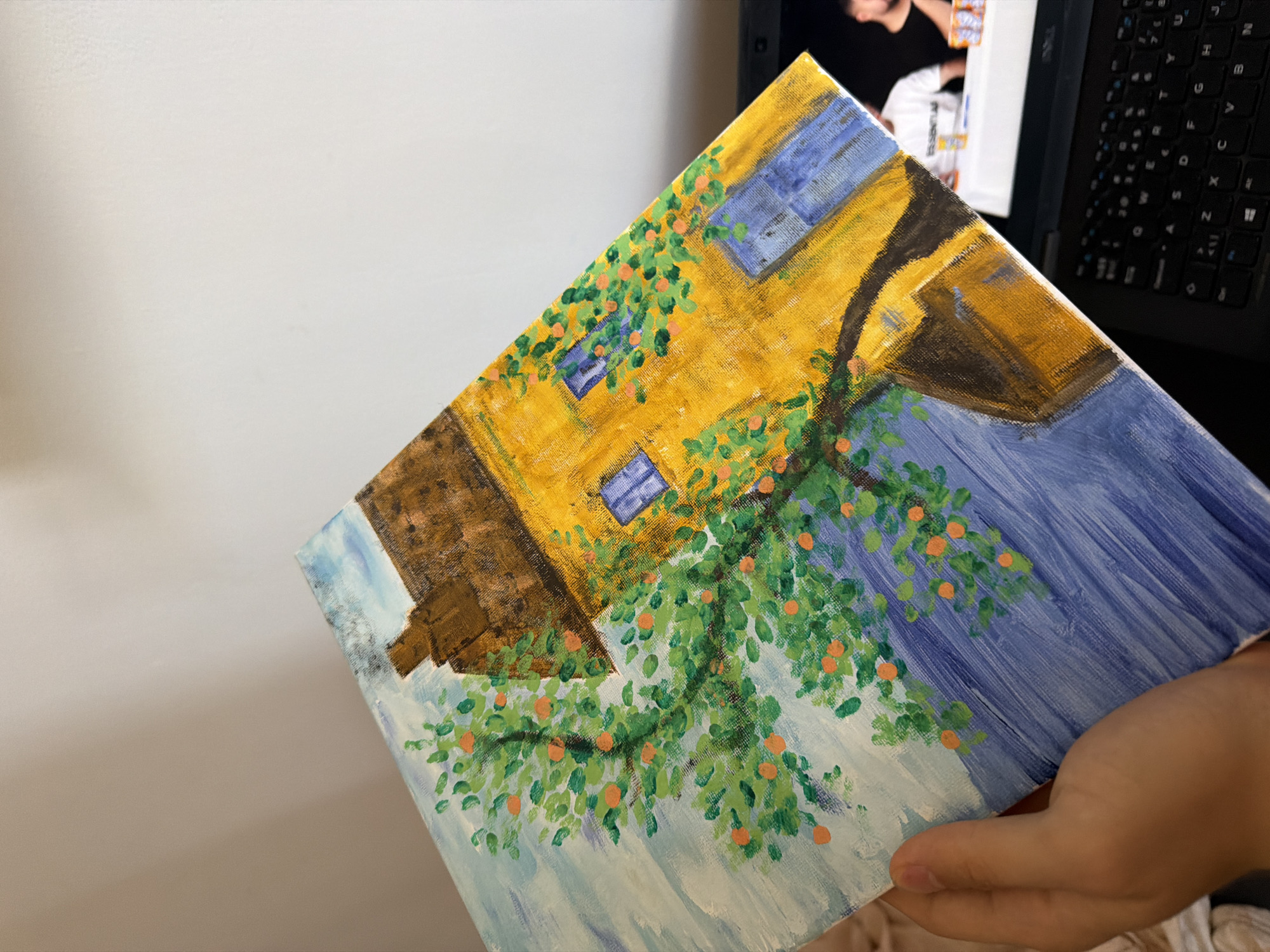 The image is a close-up, angled shot of an acrylic painting of a yellow house with blue shutters and a brown roof, situated next to a tree with green leaves and orange fruit. The painting is held by a person's hand. The background is a wall. The scene does not provide any information about the city of Sofia, Bulgaria, or any specific location.