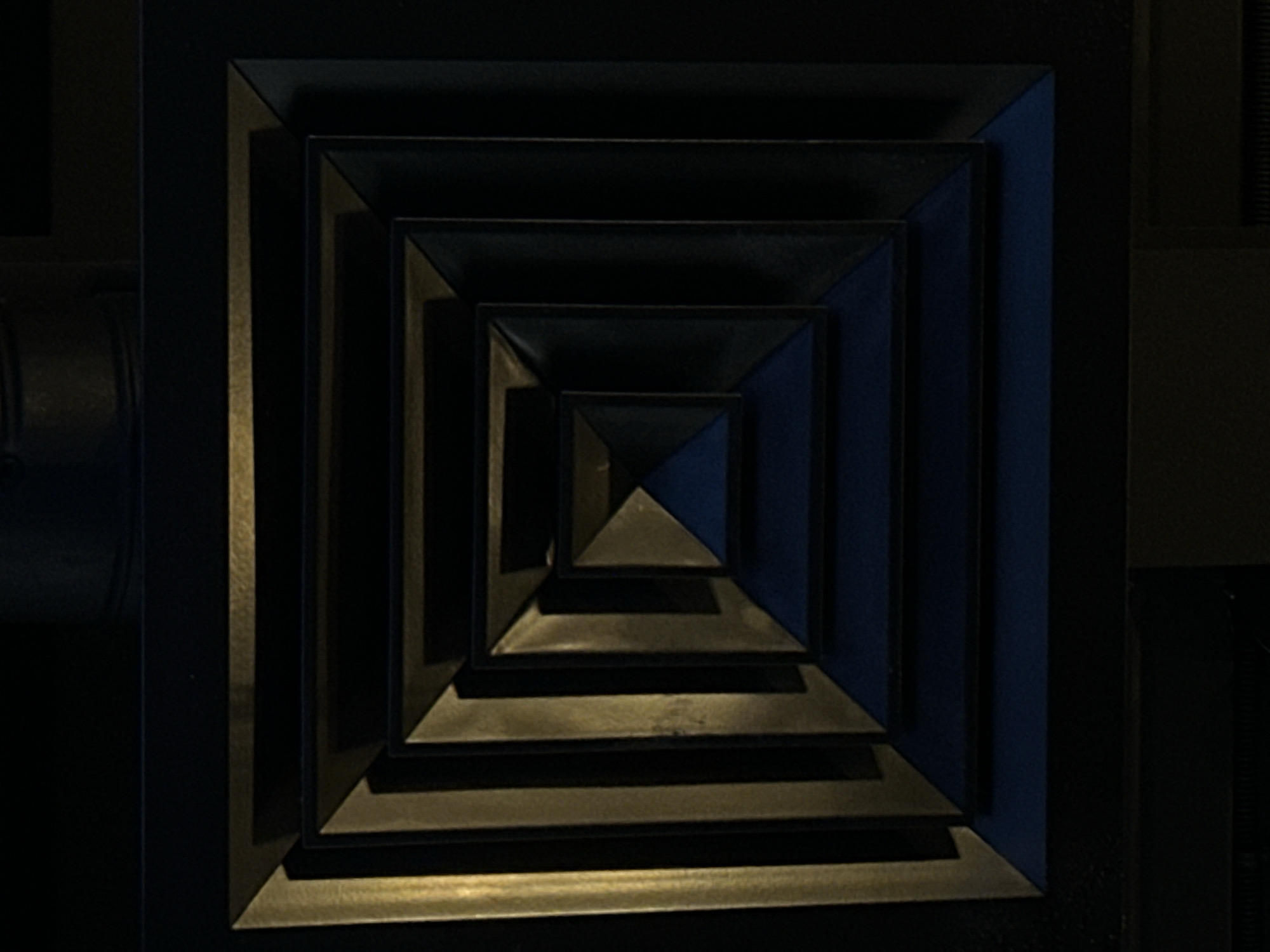 The image depicts a square vent, likely an air conditioning or ventilation duct, composed of concentric squares diminishing in size towards the center. The vent appears to be made of a dark metal, possibly steel or iron, with subtle variations in tone suggesting a dark golden or bronzed finish. It is situated on a dark, possibly black or very dark gray, background. There is limited ambient light, resulting in strong shadows that accentuate the dimensionality of the vent structure. A portion of what seems to be a cylindrical metal pipe is visible on the left edge of the frame. The location in Votkinsk, Russia is not inferable from the image.
