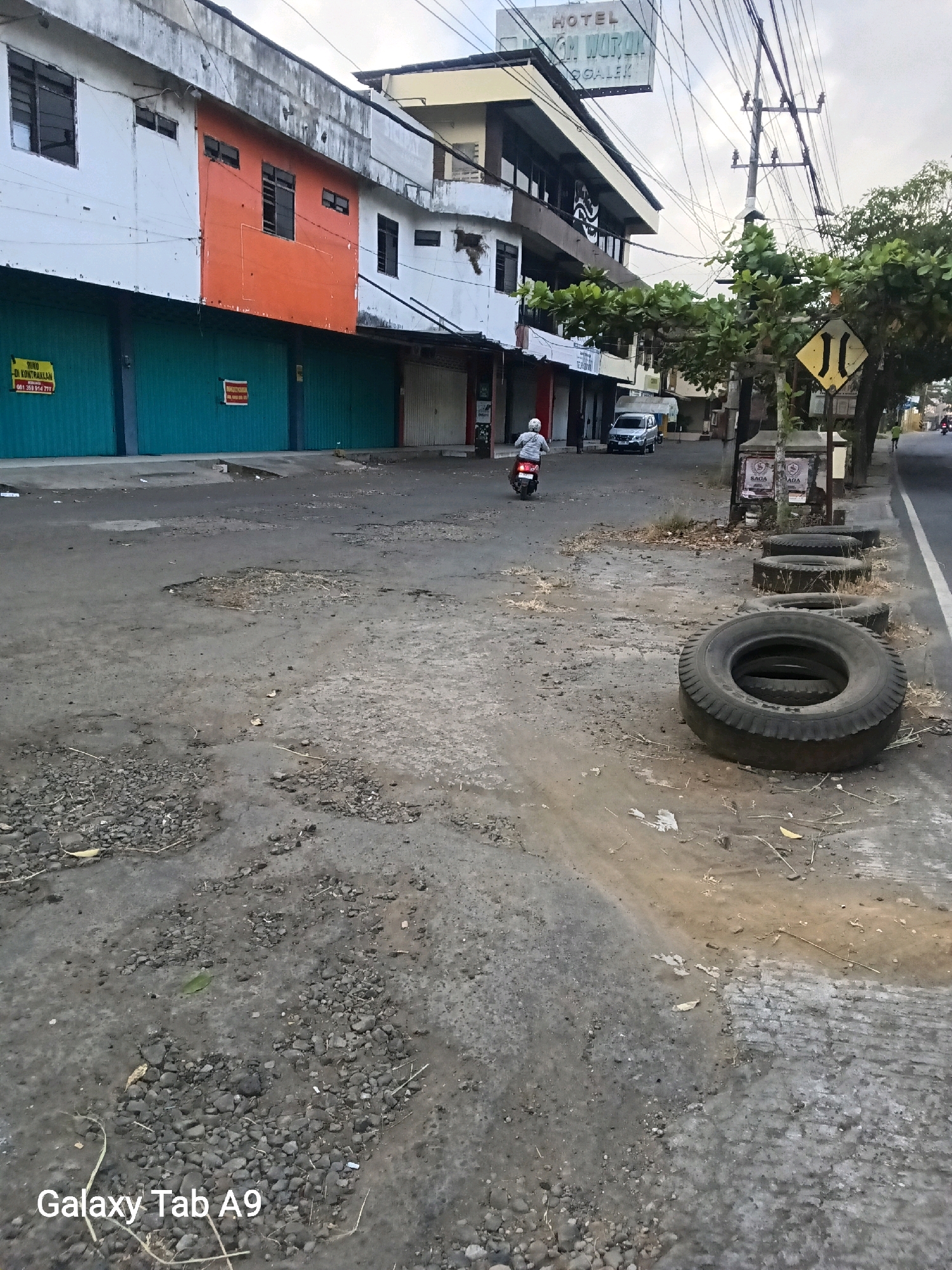 A street scene in Trenggalek, Indonesia, shows a weathered building with an orange section and turquoise shutters, a sign for "HOTEL DHARM WURAK," and a motorcycle rider. The road is cracked and has potholes, with debris scattered along the side. Several discarded tires are stacked on the roadside next to a traffic sign indicating a narrow road. Trees and power lines are visible, and the sky is overcast.