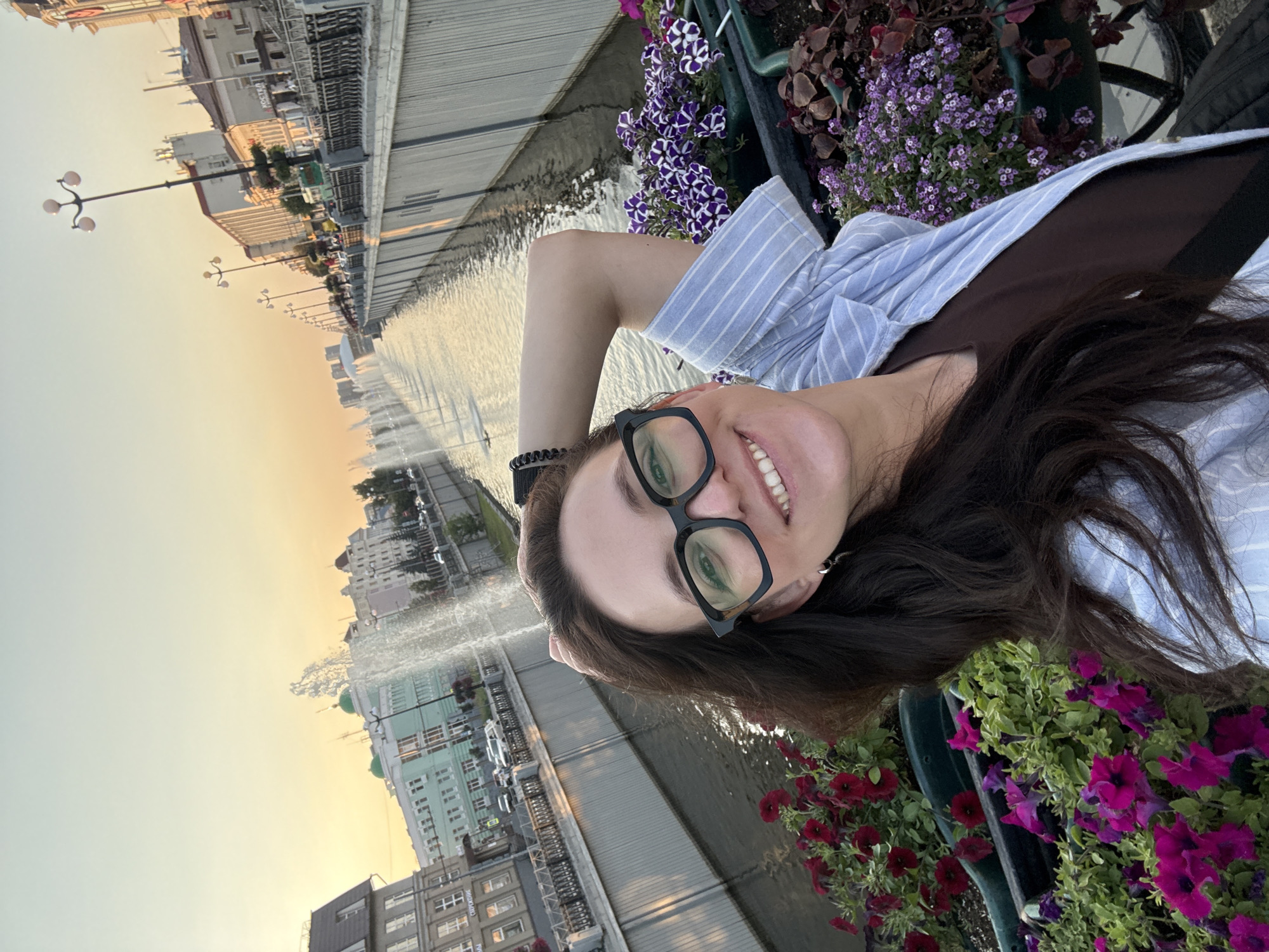 A woman takes a selfie on a bridge in Kazan, Russia, with fountains and buildings in the background. She is wearing glasses and a blue and white striped shirt. She is smiling at the camera with her arm behind her head. In the foreground, there are planters filled with pink and purple flowers. The sky is a soft orange and yellow, indicating either sunrise or sunset. The architecture in the background suggests a historic city center.