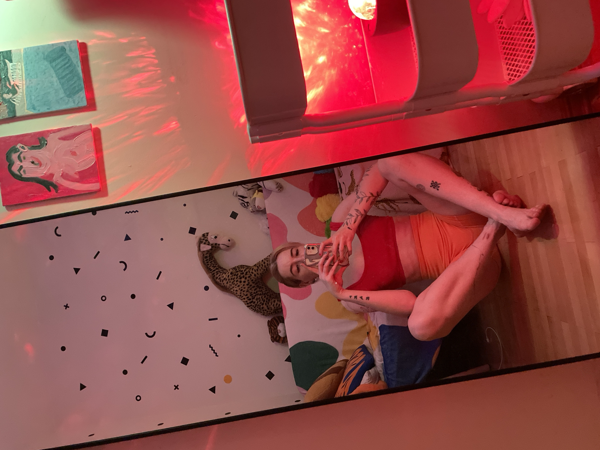 A full-length, medium shot depicts a woman lying on her side on a bed in a room with a red light. The woman is wearing a red top and orange shorts and is holding a phone in her hand, presumably taking a picture. Her legs are bent at the knees. Tattoos are visible on her arms and legs. The bed has a colorful patterned comforter and is littered with various pillows and a stuffed animal. The wall behind the bed is decorated with a geometric pattern of black shapes on a white background. Above the bed are two small paintings and a red light fixture. The floor next to the bed is wood. The location is a room in a city, potentially Belgrade, Serbia.