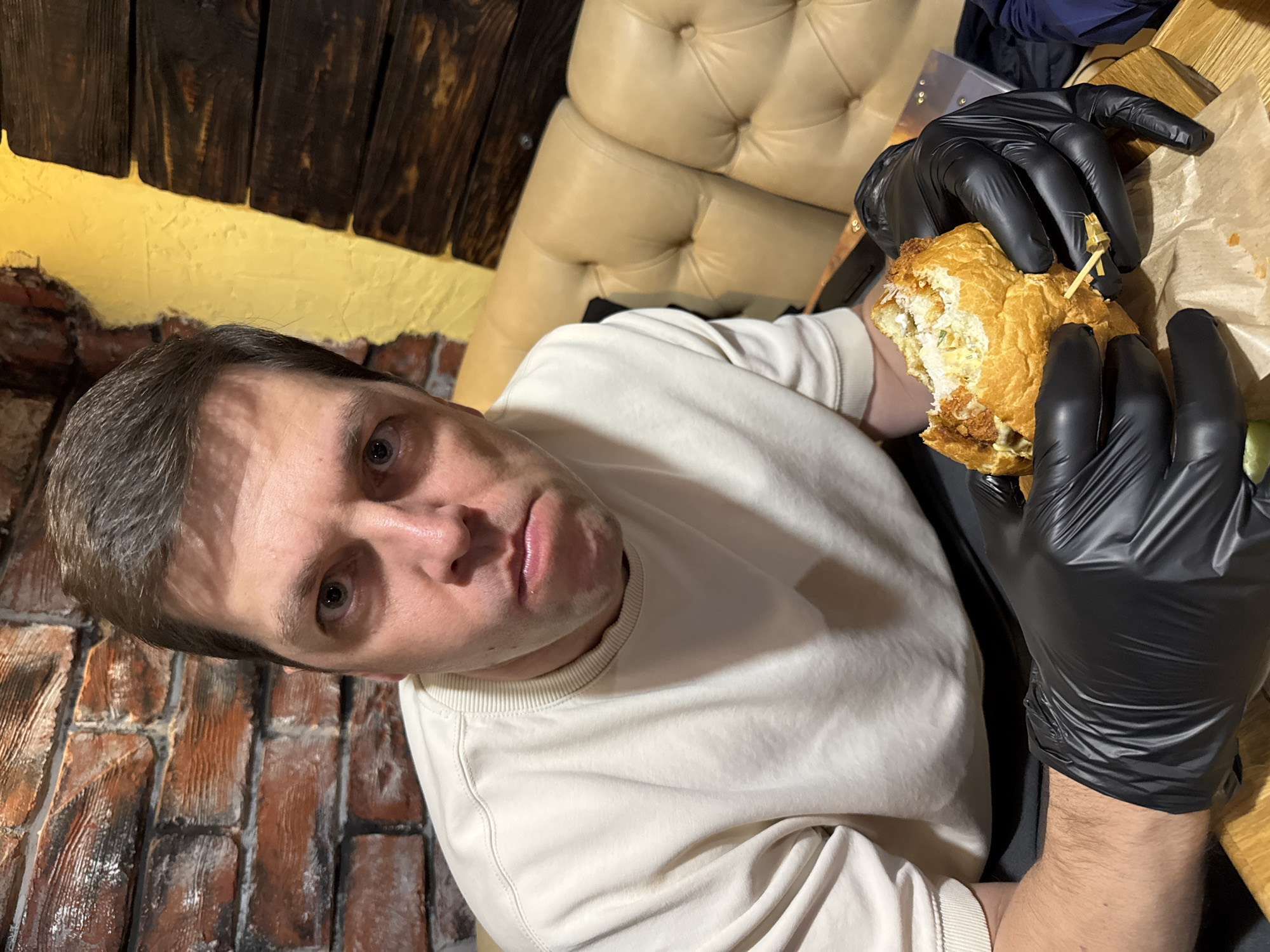 A medium close-up photograph shows a man holding and eating a burger in what appears to be an indoor restaurant setting. The man, with short brown hair and a neutral expression, looks directly into the camera. He is wearing a light-colored (cream or off-white) long-sleeved sweatshirt. Both his hands are covered in black disposable gloves as he holds a substantial, partially eaten burger, which appears to be held together by a small wooden skewer. Inside the burger, fried components, sauces, and greens are visible.
The background elements suggest a rustic and cozy interior. A reddish-brown brick wall is visible on the left side of the frame, transitioning upwards to a vibrant yellow painted wall section. Above this, dark wooden planks or beams are visible, possibly forming a ceiling or upper wall. To the man's right and behind him, a plush, beige tufted booth or seating provides a seating area. The lighting is warm and artificial, characteristic of an indoor evening or night setting. The photograph is notably rotated clockwise, positioning the man's head on the left and his body horizontally across the frame. No visible text is present.