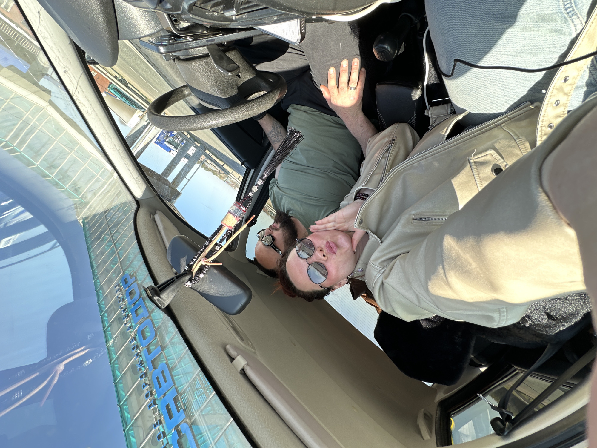 A selfie-style photo shows two people inside a vehicle. One person is seated behind the steering wheel and wearing a green shirt and sunglasses. The other person is in the passenger seat wearing a light-colored jacket and sunglasses and is turned towards the camera. Outside of the vehicle's windshield, a blue sign with Russian text and Cyrillic characters is visible. The name of the city, Ижевск (Izhevsk), Russia is written on the blue sign.