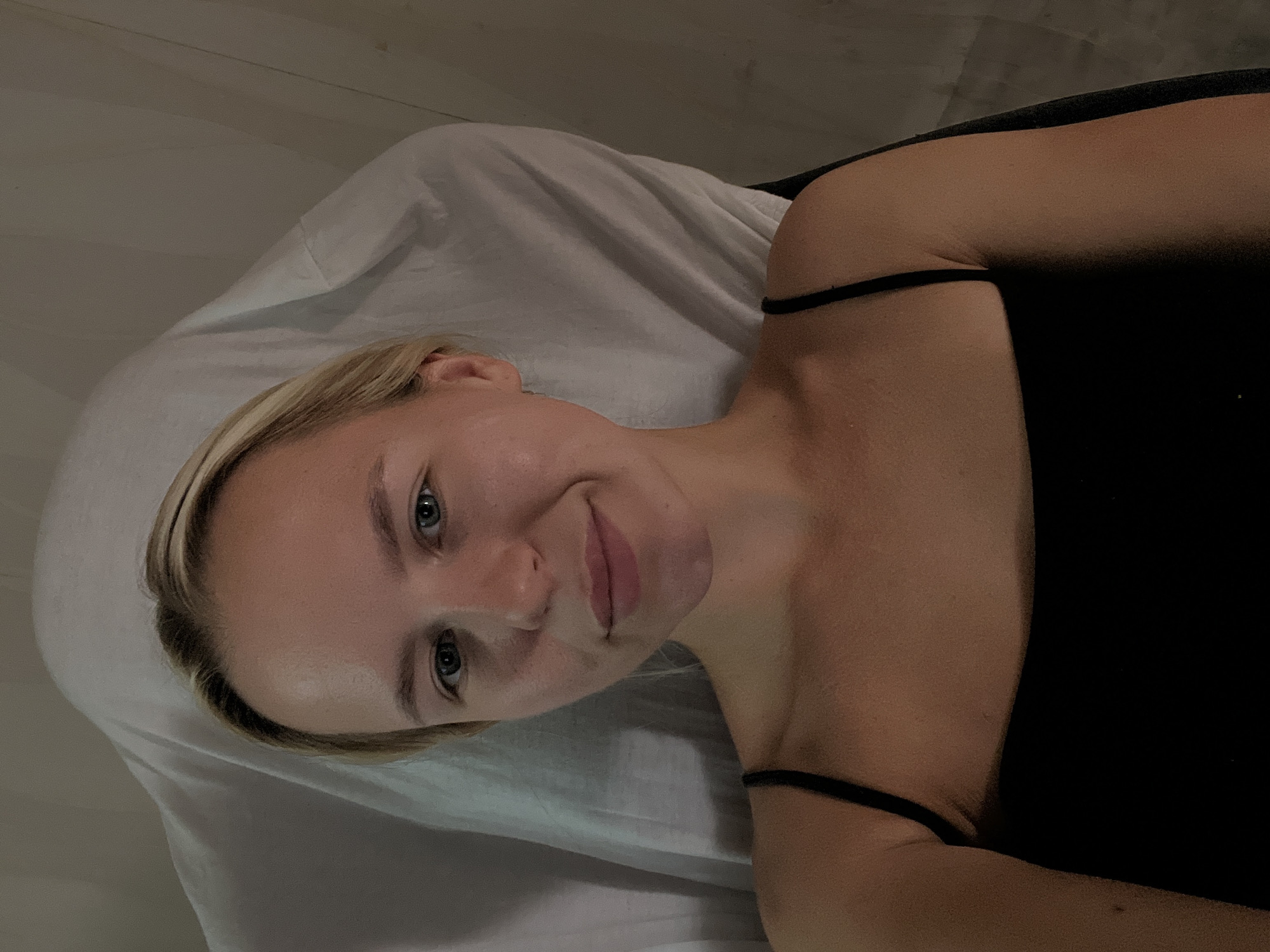 A young woman with blonde hair is lying down, looking at the camera. She is wearing a black tank top. The background is a white sheet and a dark wall. The lighting is dim and warm. There are no other people or objects visible in the image.