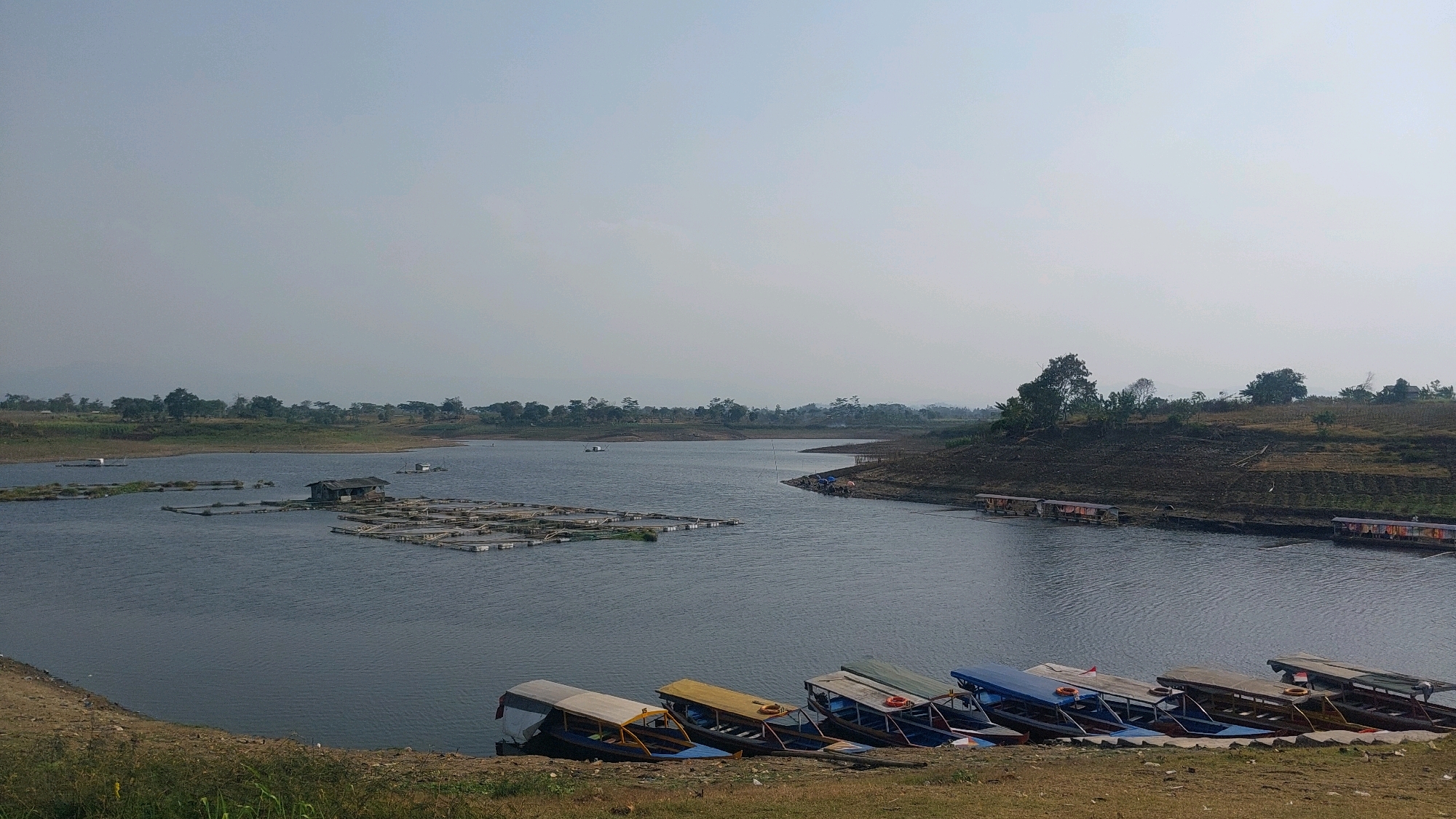 The image depicts a serene reservoir in Bojonghaleuang, Indonesia, under a hazy, pale sky. The water's surface is calm, with gentle ripples indicating a light breeze. In the foreground, a line of colorful boats, primarily yellow, blue, and white, are moored along a dirt embankment. These boats appear to be passenger vessels, possibly used for tours or transportation across the reservoir.
Further out on the water, a collection of floating structures, likely fish cages or aquaculture farms, are visible. One of these structures features a small, rustic hut. In the distance, the reservoir stretches towards a landscape characterized by rolling hills covered in sparse vegetation and scattered trees. The treeline suggests a rural or semi-rural environment. The overall impression is one of tranquility and natural beauty, with a hint of human activity through the presence of the boats and aquaculture.