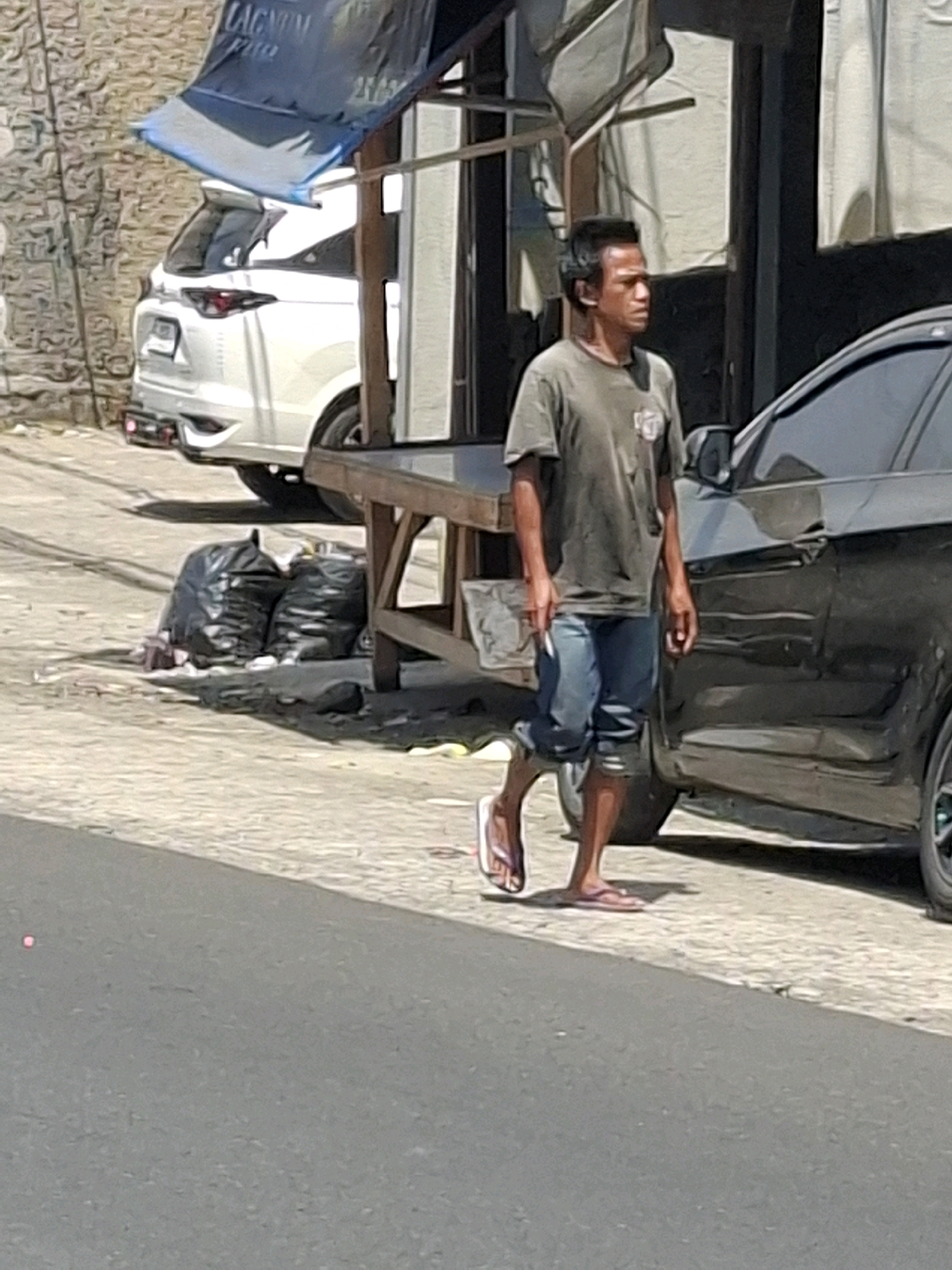 A man is walking on the sidewalk next to a black car. He is wearing a green t-shirt and blue shorts, with flip-flops on his feet. Behind him, there are two black trash bags on the ground. In the background, a white SUV is parked in front of a building. The scene appears to be on a street in Cikahuripan, Indonesia, with a brick wall and a stall structure visible. The weather is sunny, casting shadows on the ground.