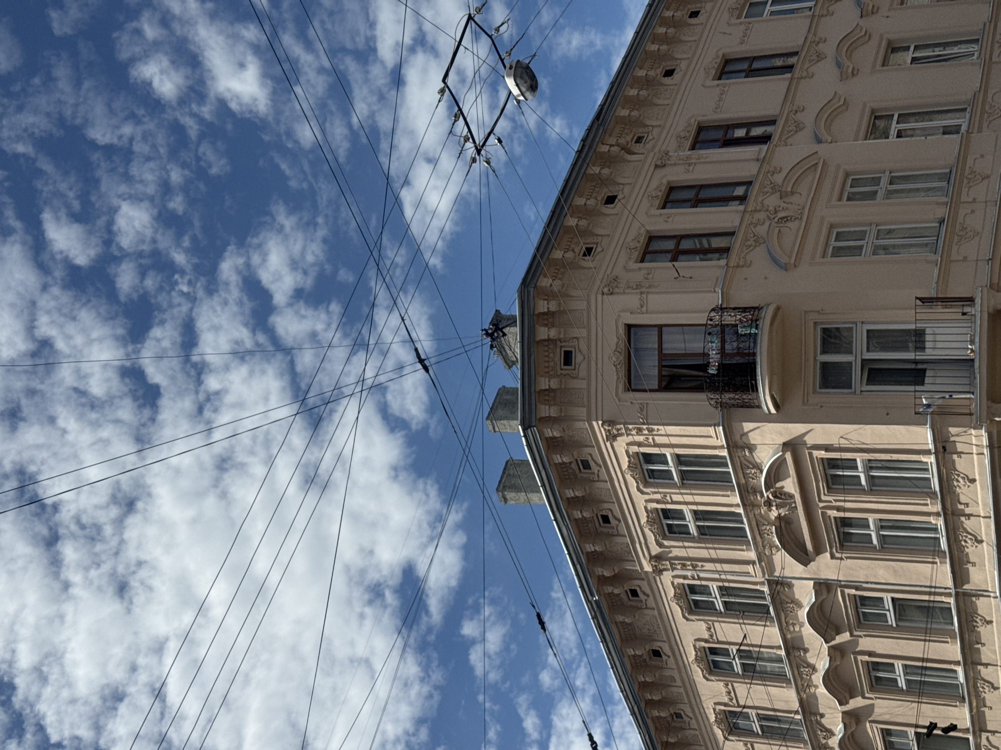 A low-angle, vertical medium shot captures the corner of a light-peach building with ornate architectural details under a bright blue sky dotted with fluffy white clouds. A complex network of dark overhead wires crisscrosses the sky, with a trolleybus contact wire assembly visible in the upper right quadrant. The building features multiple rows of windows with white frames and dark panes, some of which are open. Decorative elements, including sculpted reliefs and arched window surrounds, adorn the facade. Two chimneys rise from the roofline. The scene is devoid of people.